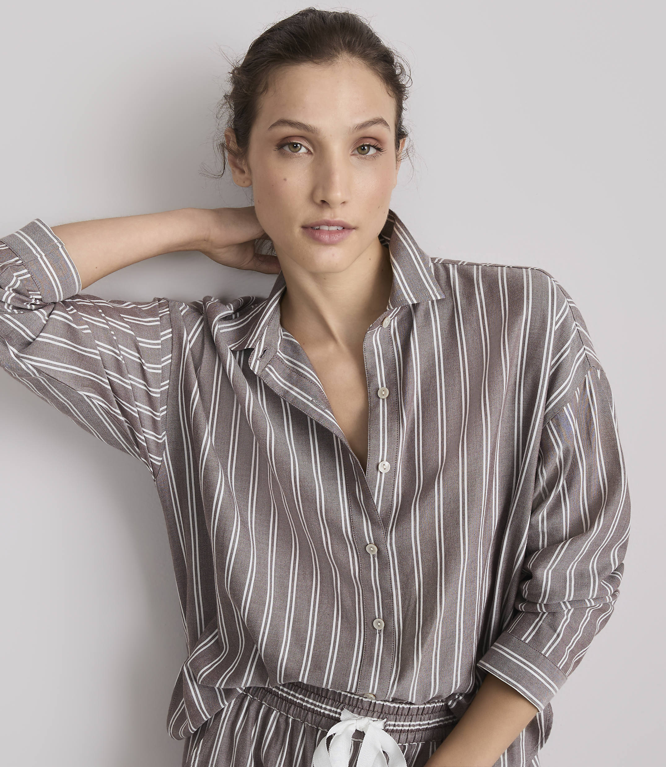 A woman in a striped pajama top rests her hand behind her head against a plain gray background.
