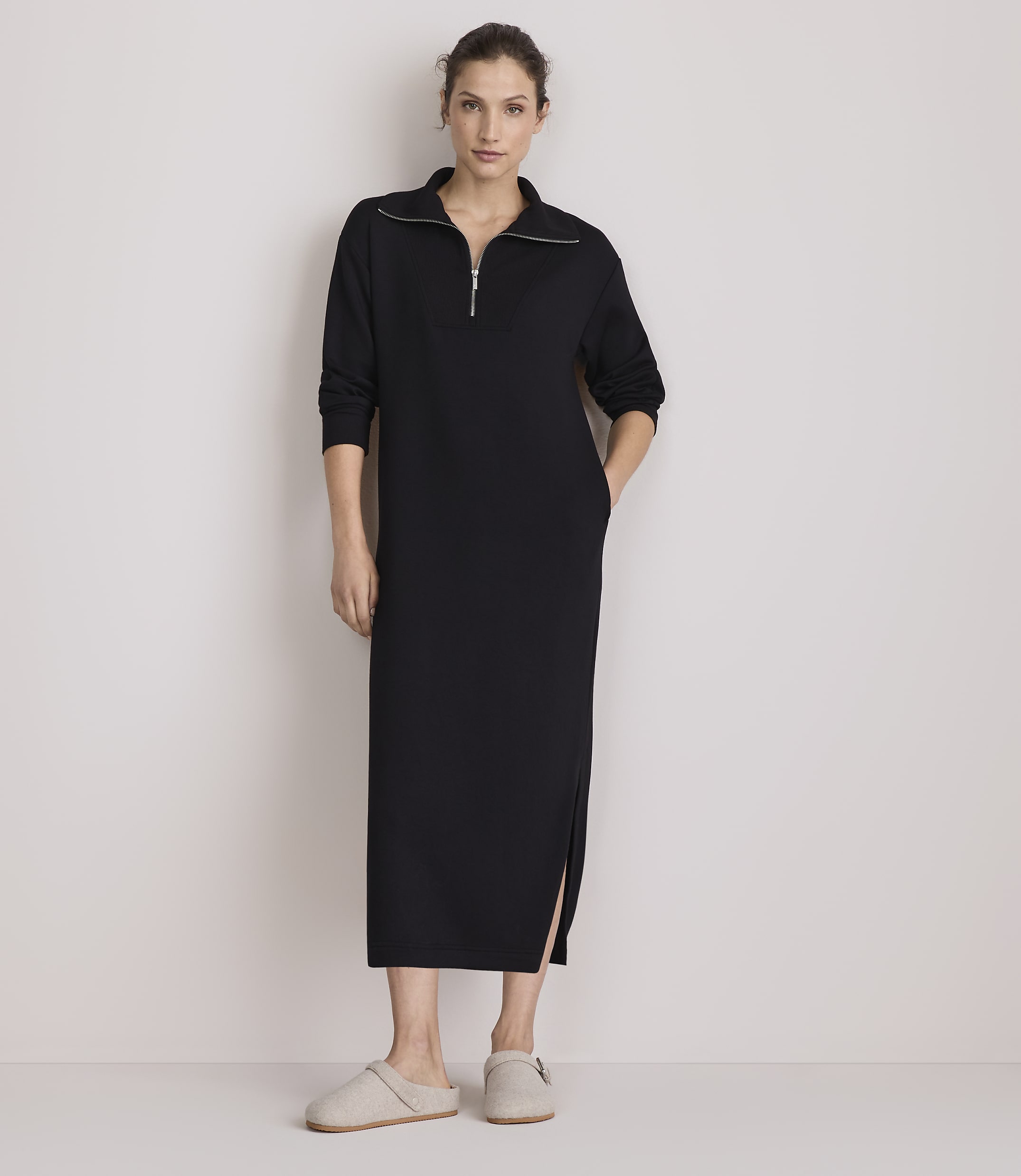A woman in a black zip-neck dress and slippers stands against a plain, light-colored wall.
