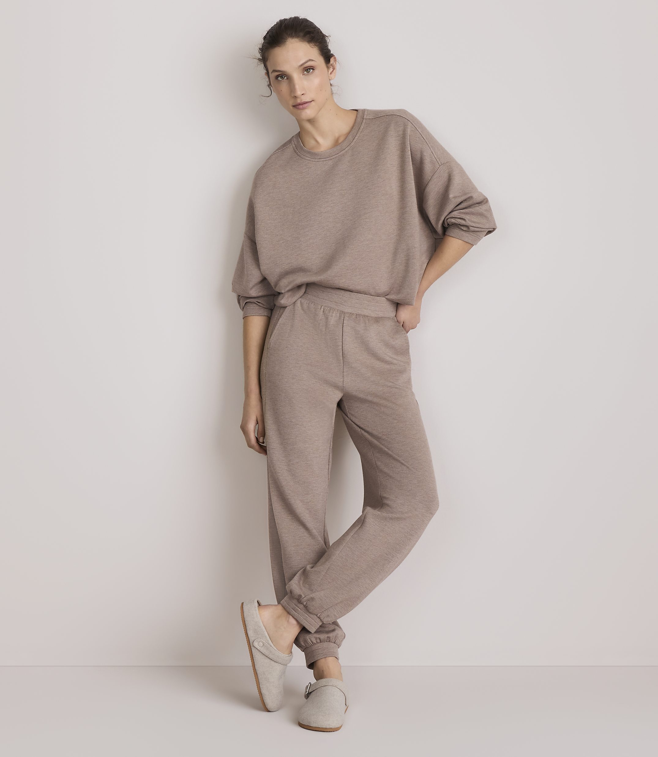 A woman in a beige loungewear set and slippers stands against a plain, light-colored wall.