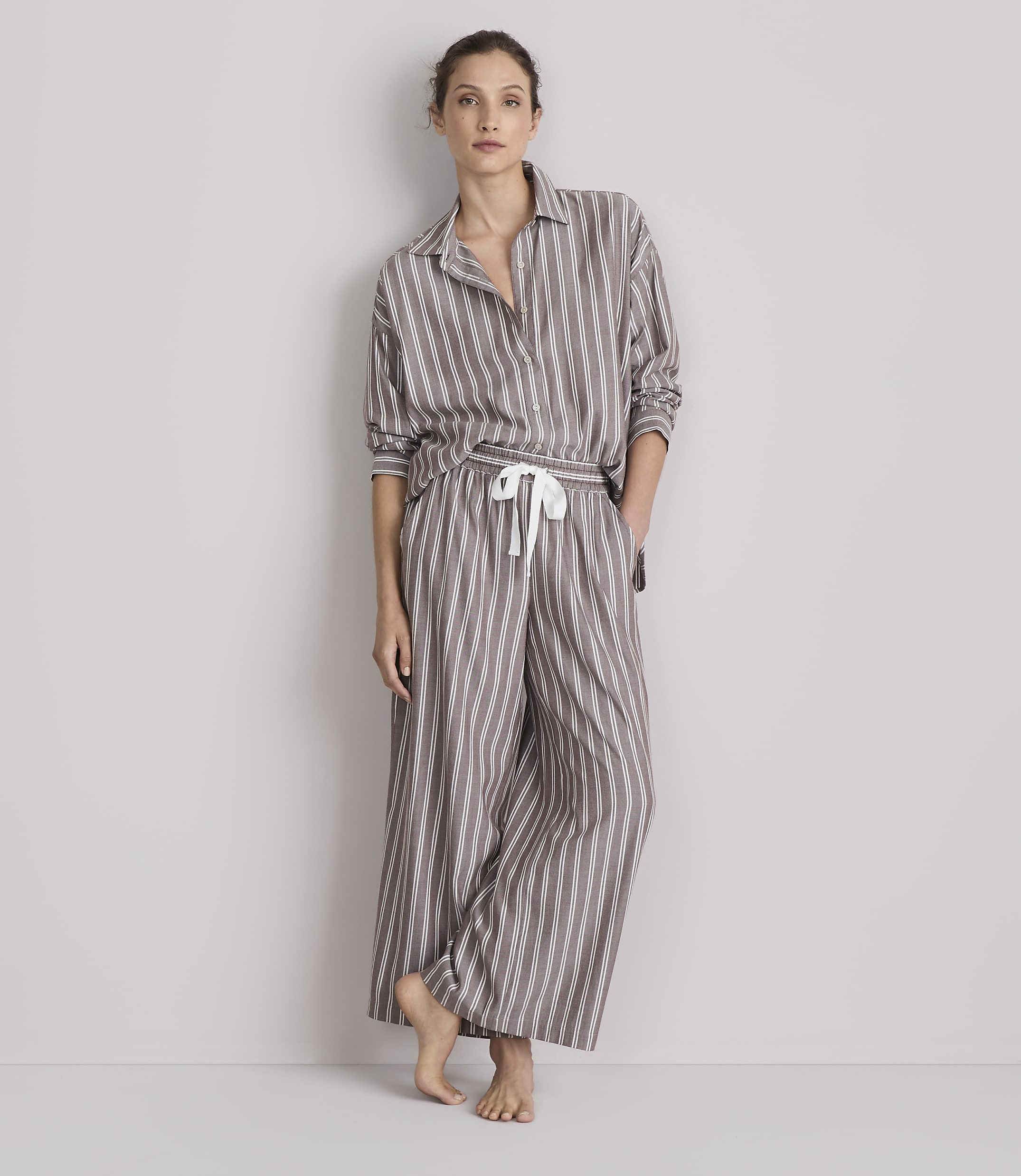 A woman stands barefoot in striped pajamas against a plain gray background.