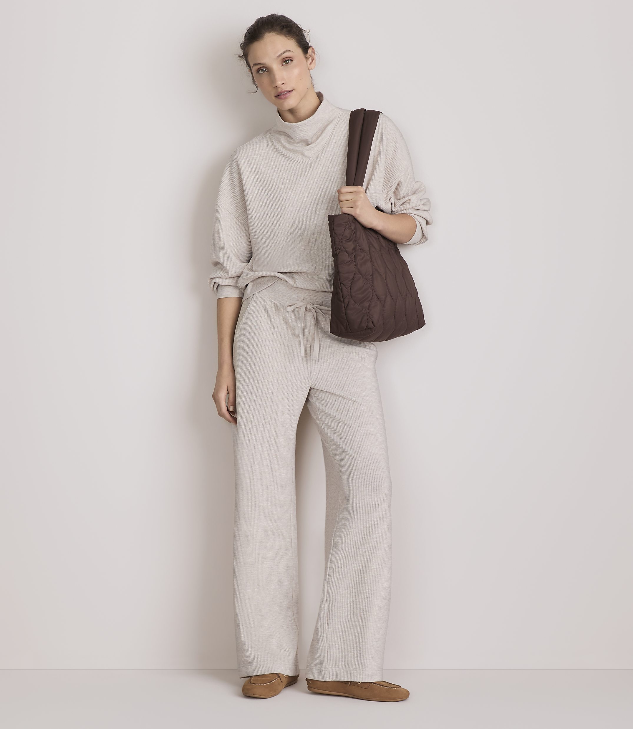 A woman in a beige loungewear set and brown tote bag stands against a plain, light-colored wall.