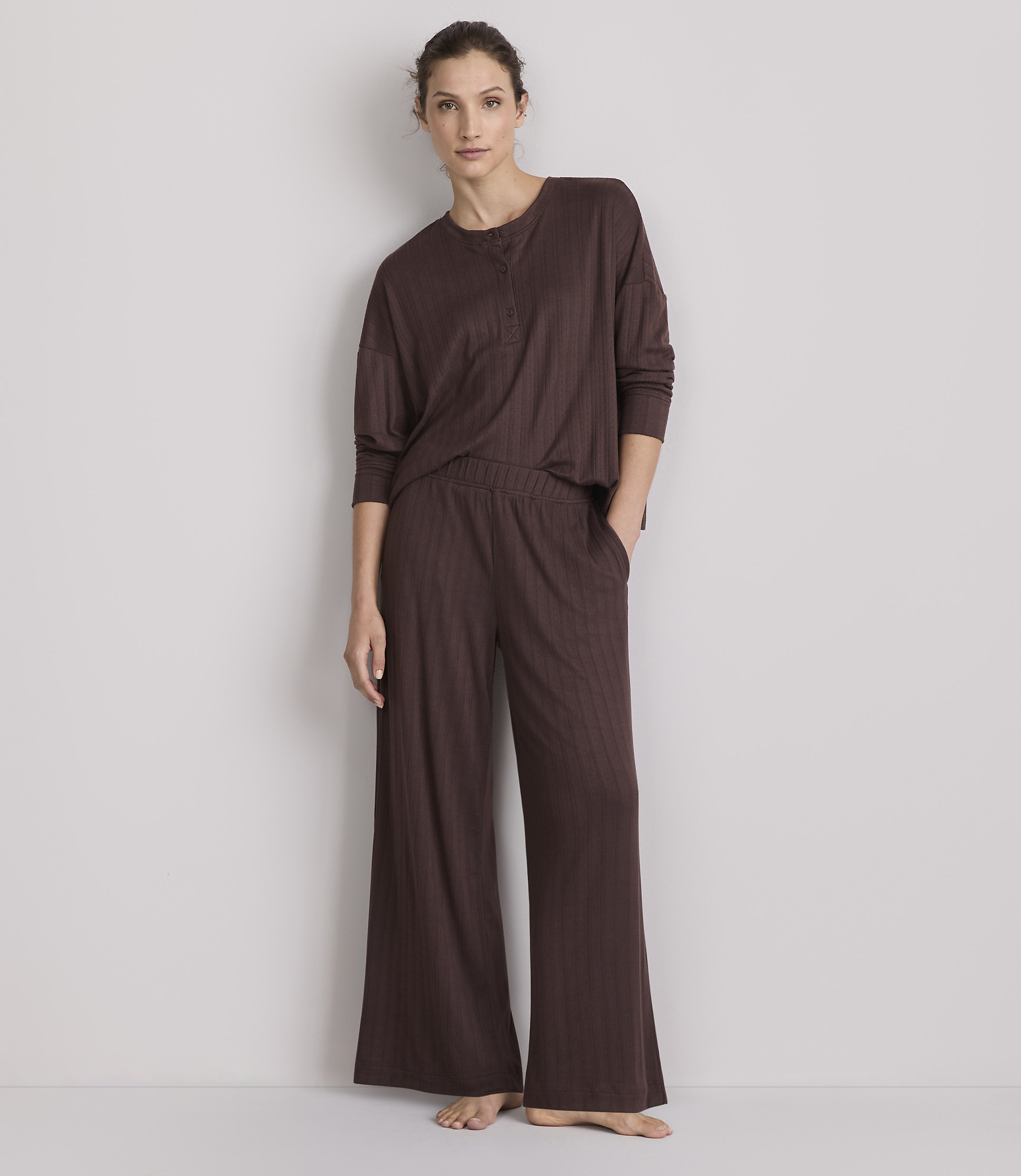 A woman in a brown ribbed pajama set stands against a plain, light-colored wall.
