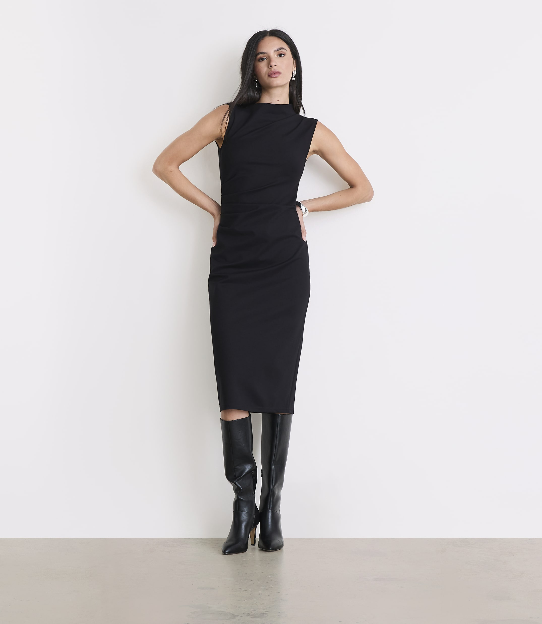 A woman in a black dress and boots stands against a plain white background.