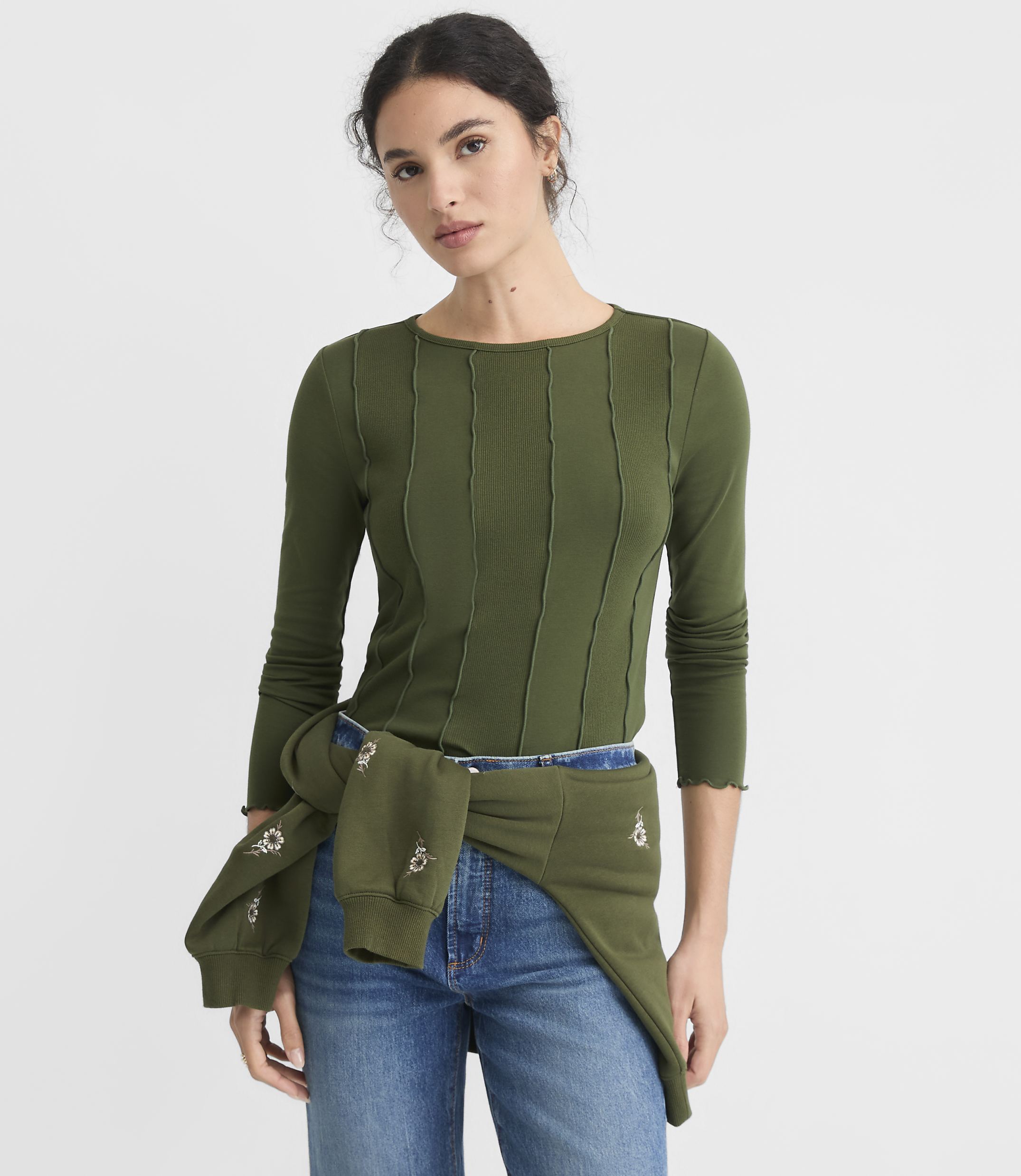 A young woman in a green ribbed long-sleeved shirt and jeans stands against a plain white background, with a green sweatshirt tied around her waist.