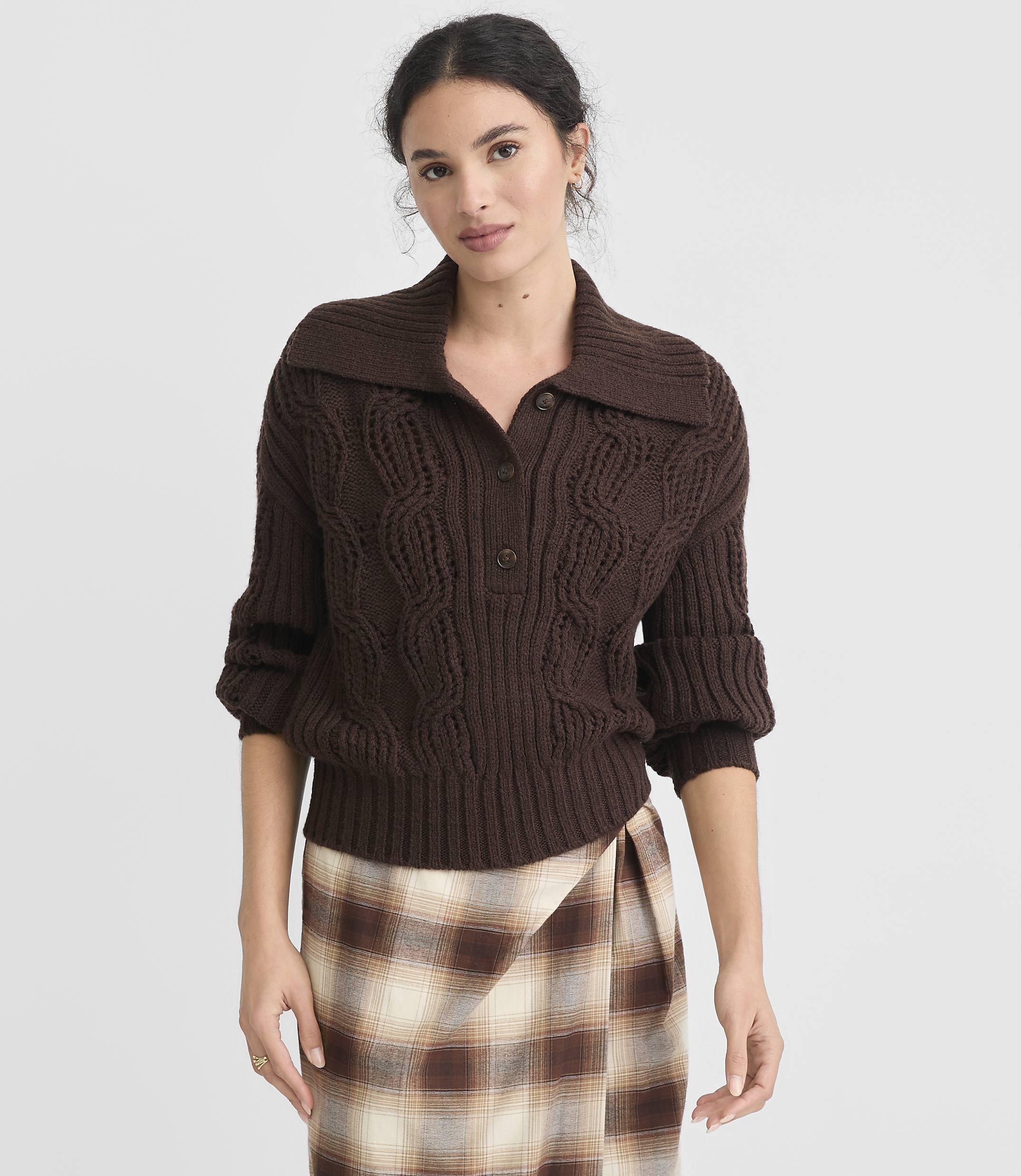 A woman in a brown cable-knit sweater and plaid skirt stands against a plain white background.