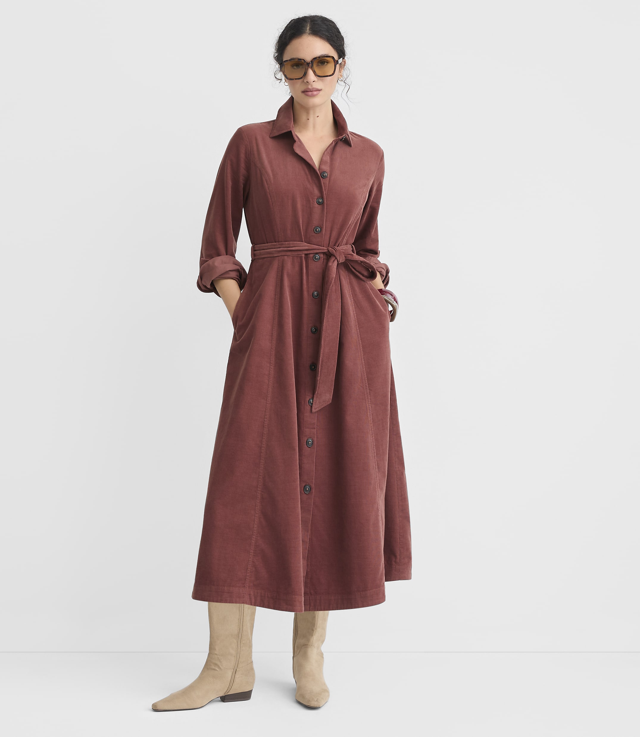 A woman in a rust-colored corduroy shirtdress with a belted waist and beige boots stands against a plain white background.