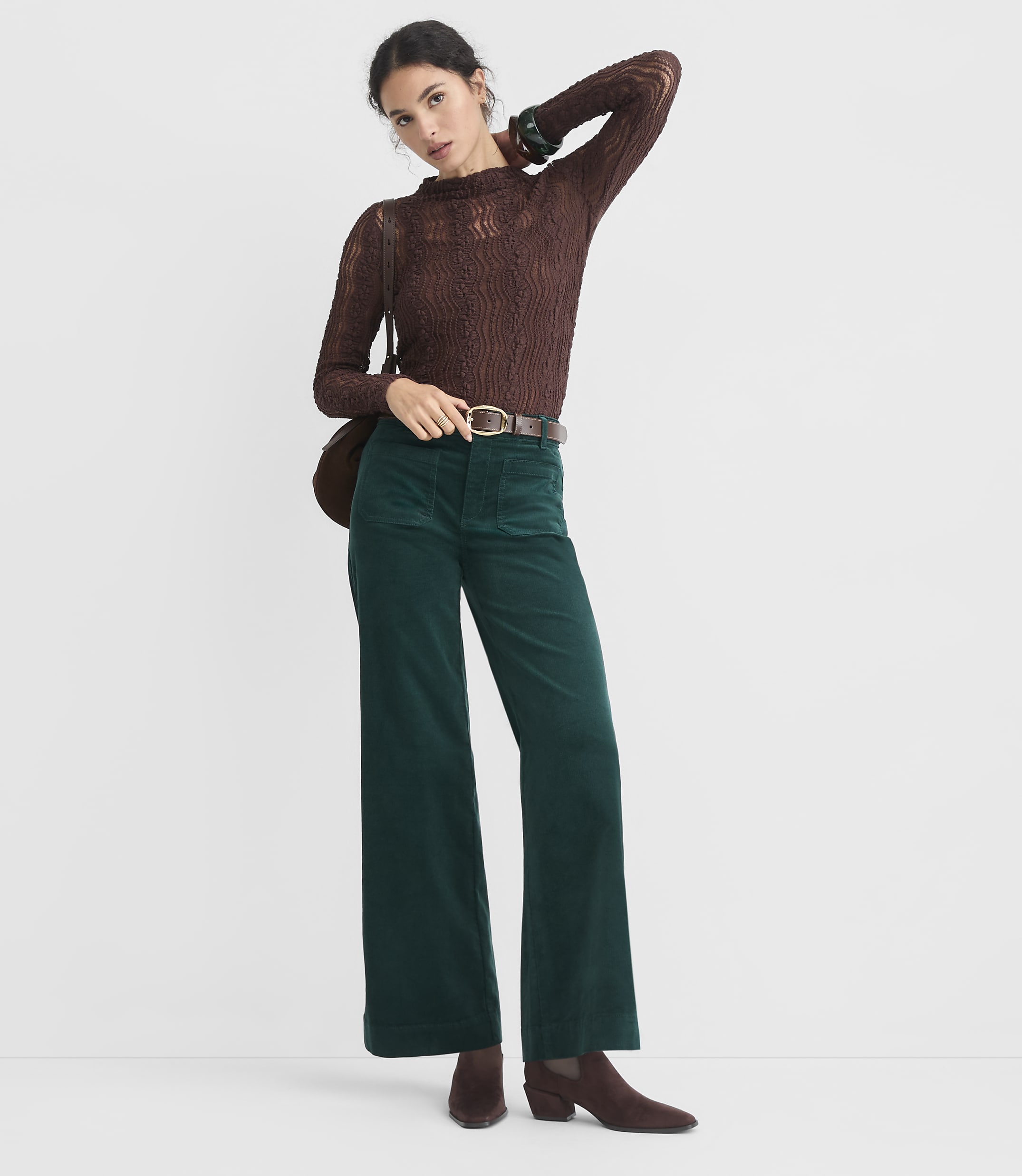A woman in a brown lace top and green corduroy pants stands against a plain white background.