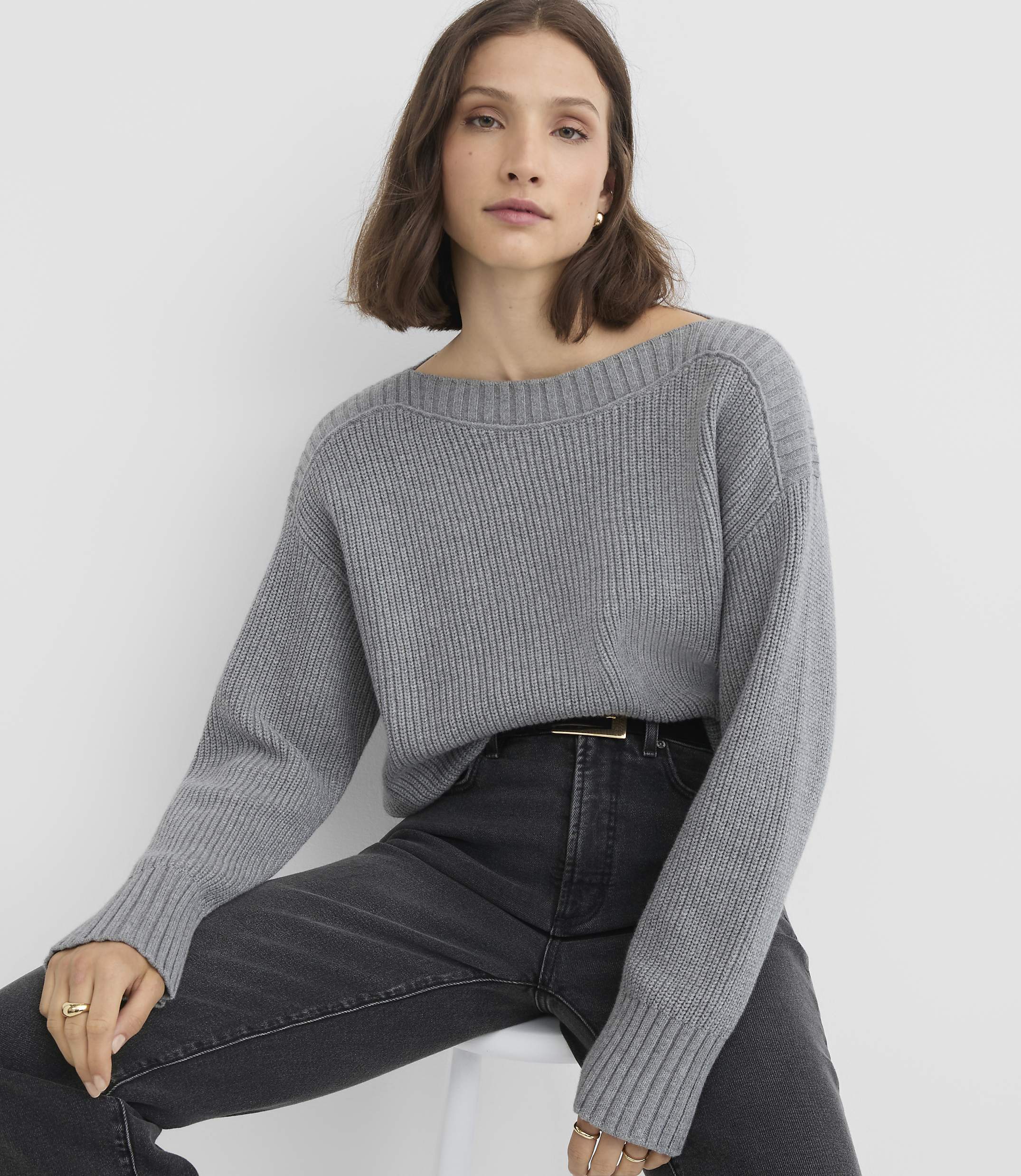 A woman in a grey sweater and dark jeans sits against a plain white background.