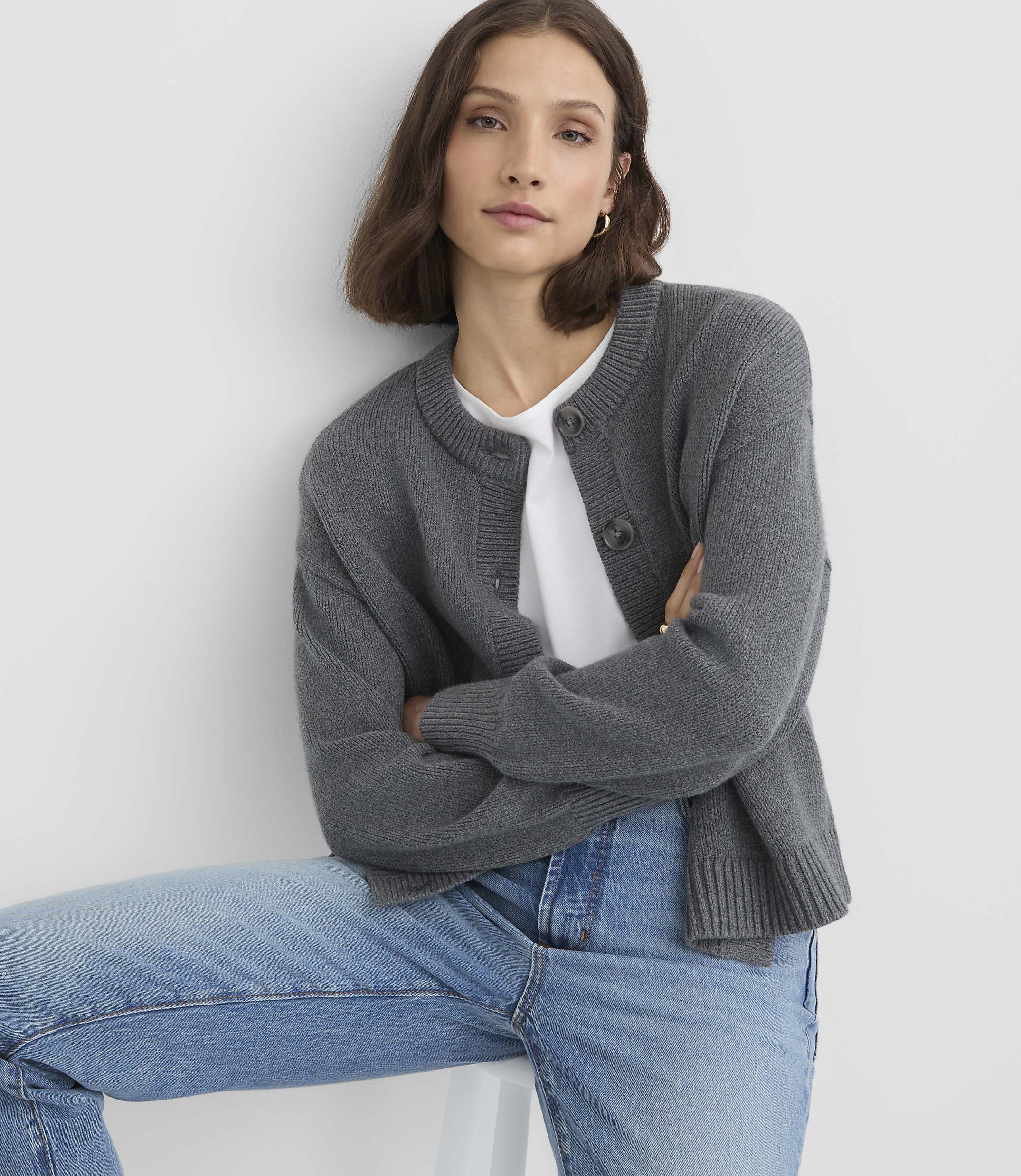 A woman in a gray cardigan and jeans sits in front of a plain white background.
