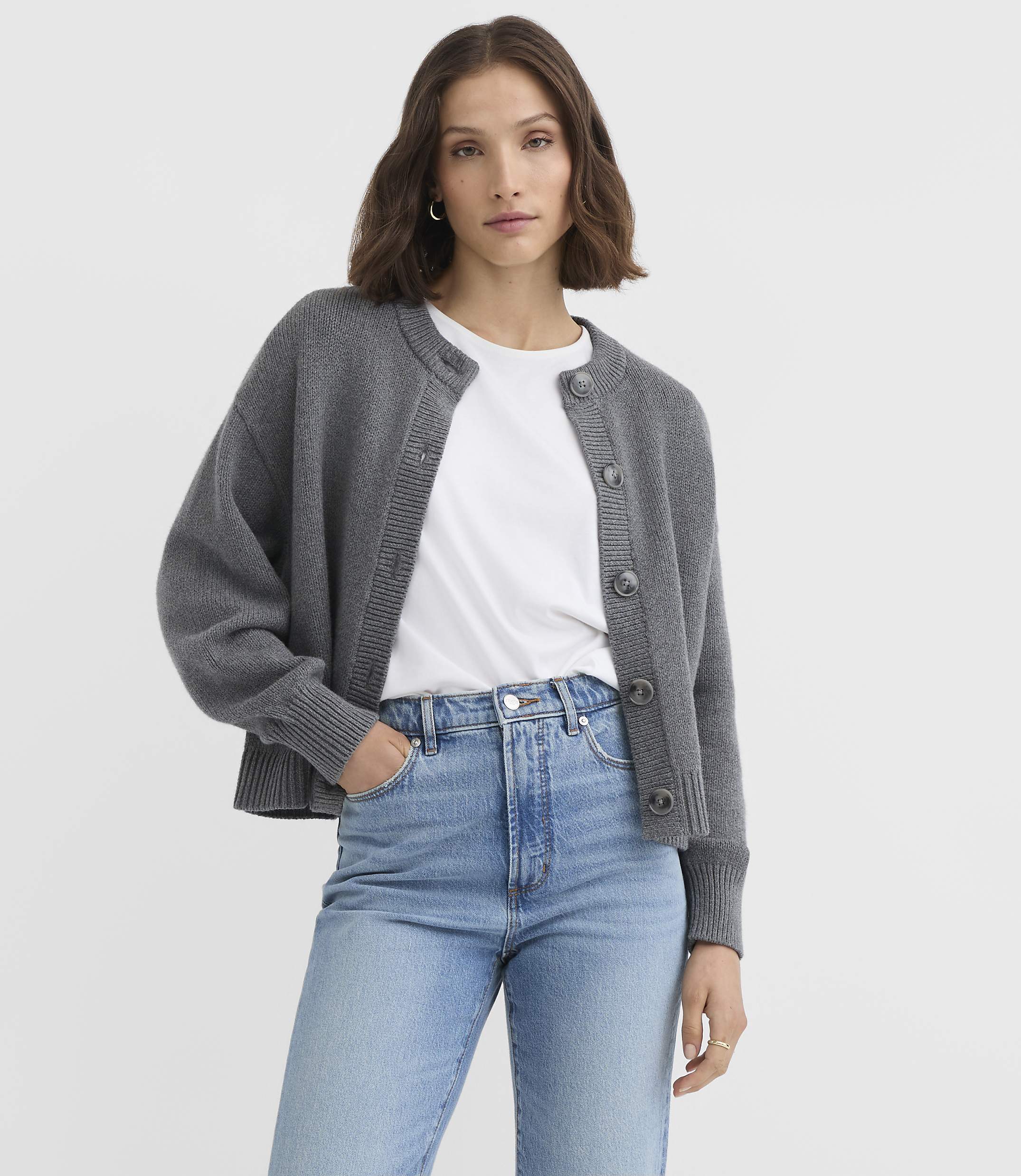 A woman in a grey cardigan and blue jeans stands against a plain grey background.