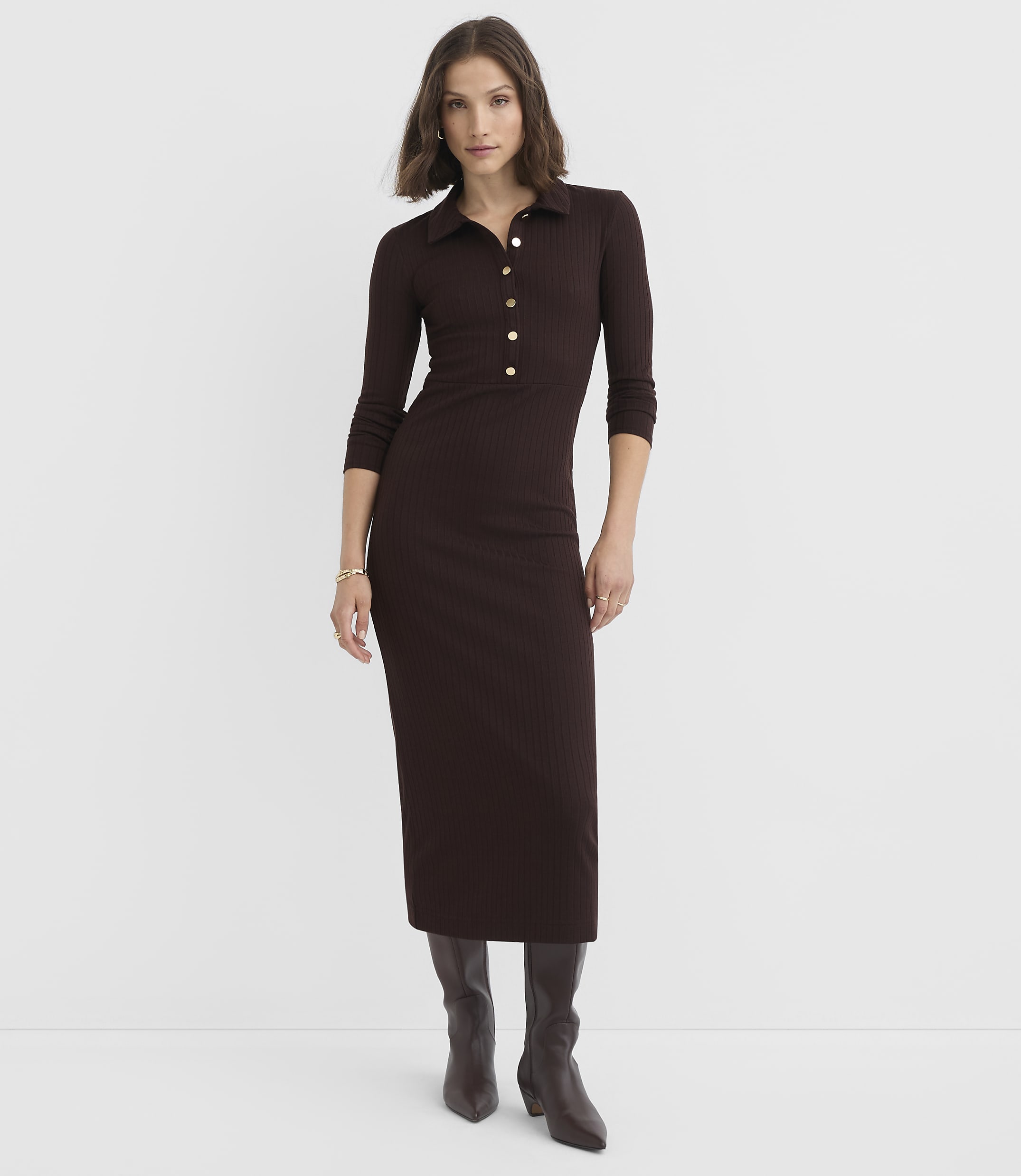 A woman in a brown ribbed midi dress and boots stands against a plain white background.