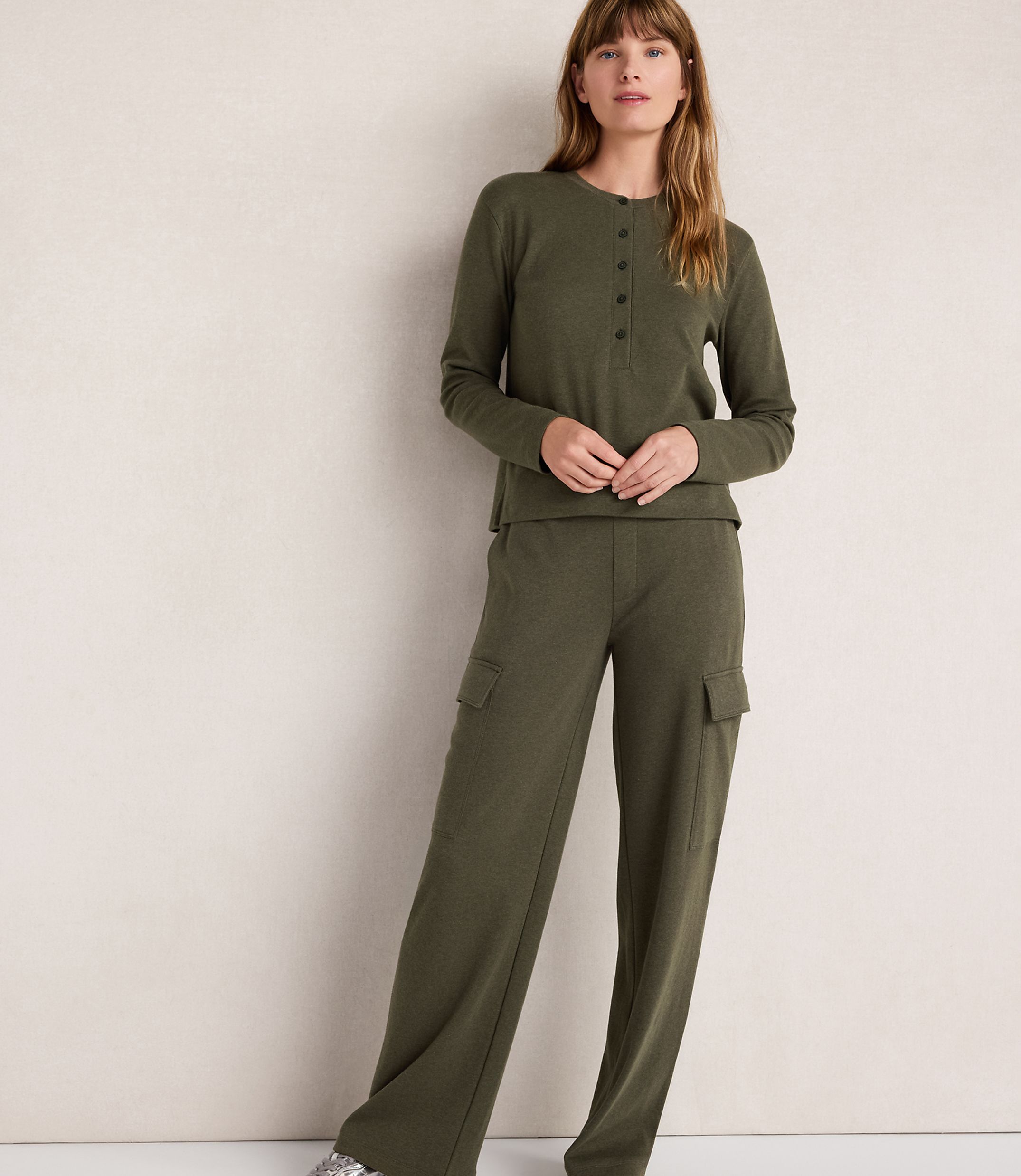 A woman in a green henley and cargo pants stands against a plain, light-colored wall.