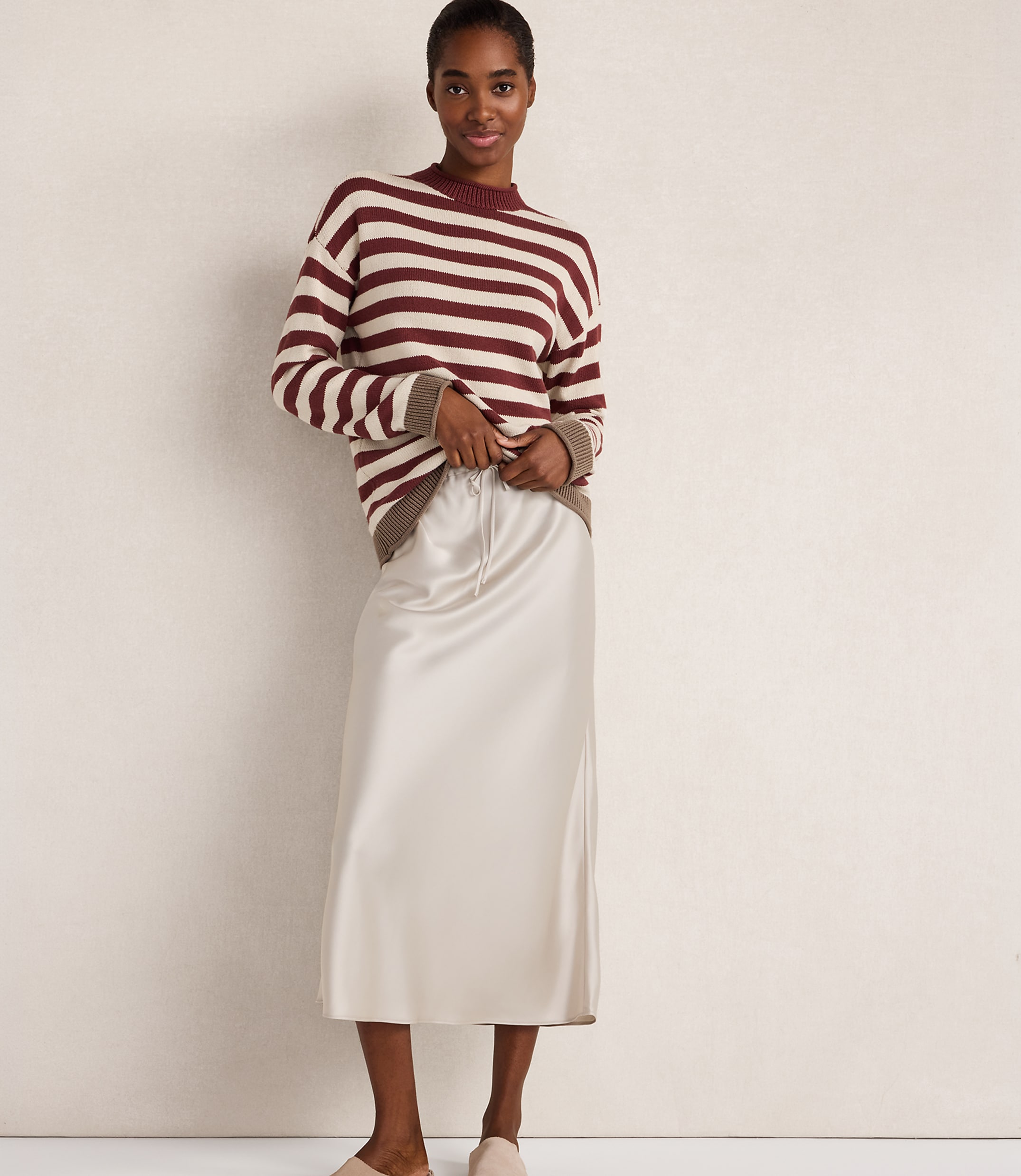 A woman in a striped sweater and satin skirt stands against a plain, light-colored wall.
