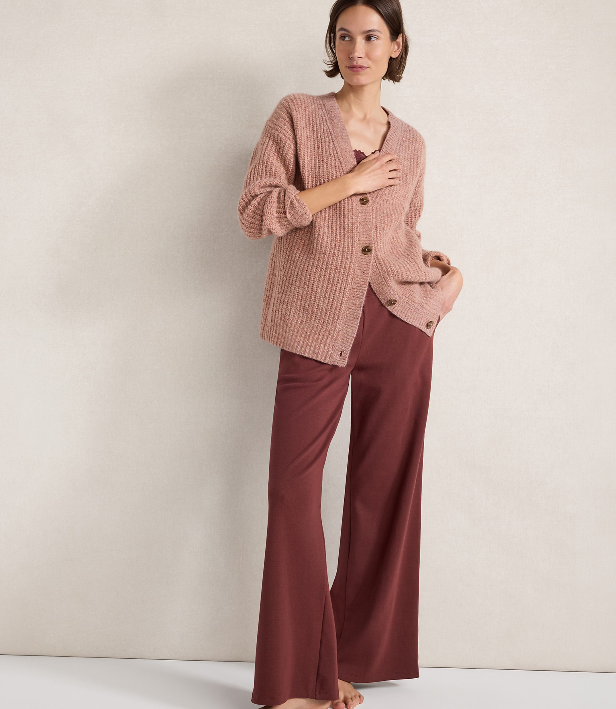 A woman in a pink cardigan and maroon pants stands against a plain, textured background.