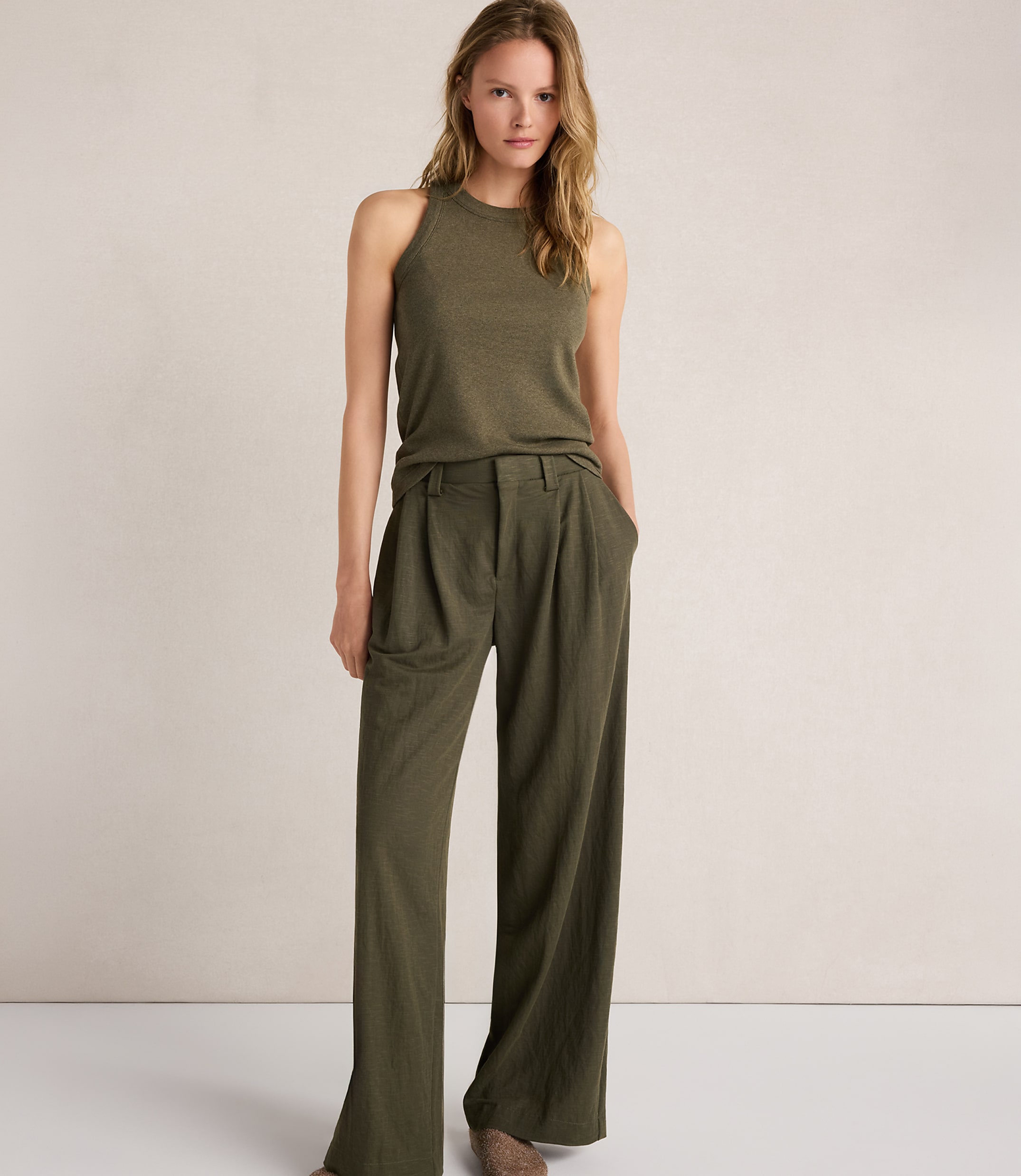 A woman in a green tank top and wide-leg pants stands against a plain, light-colored background.
