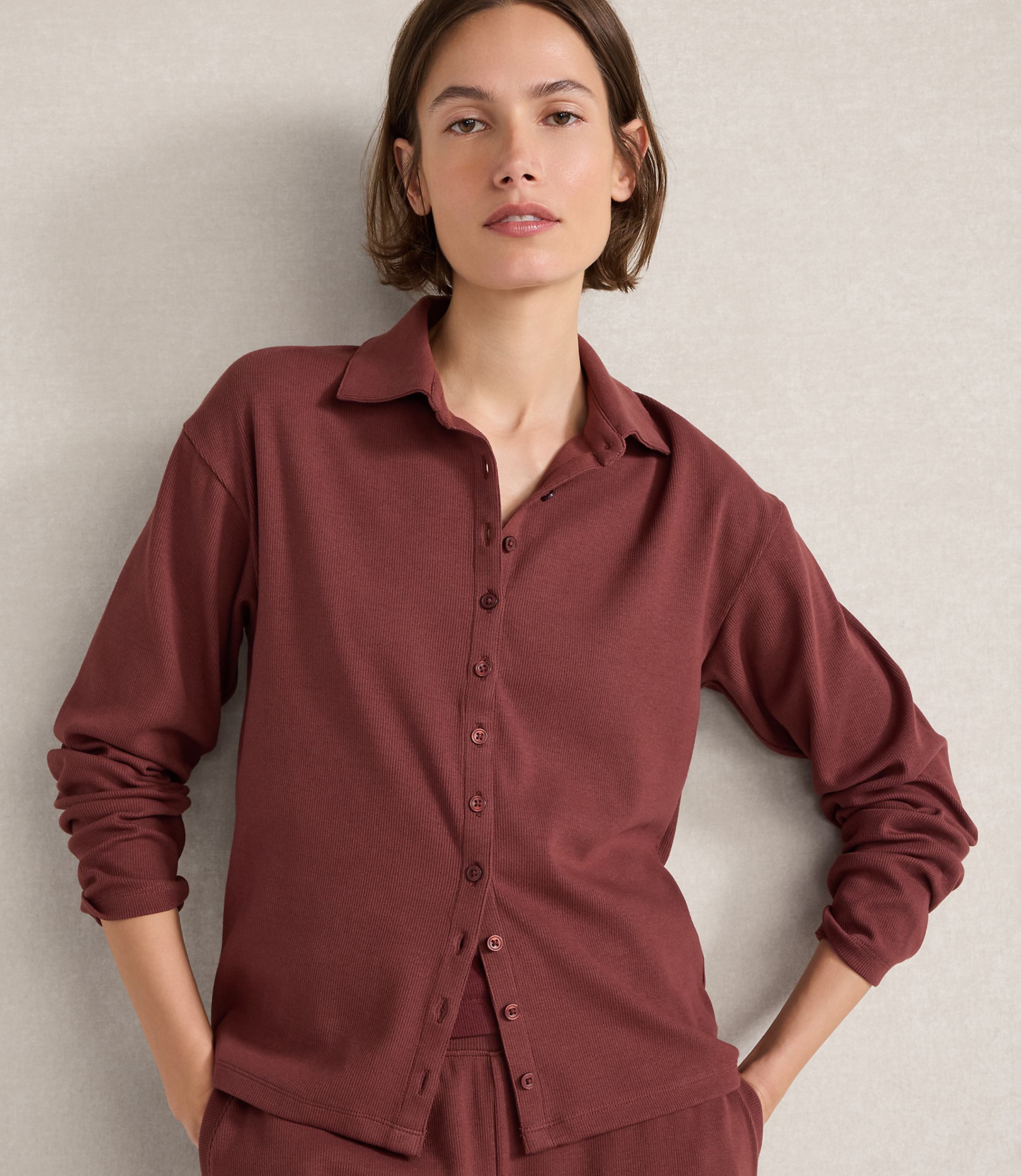 A woman in a maroon ribbed button-down shirt and matching pants stands against a textured gray wall.