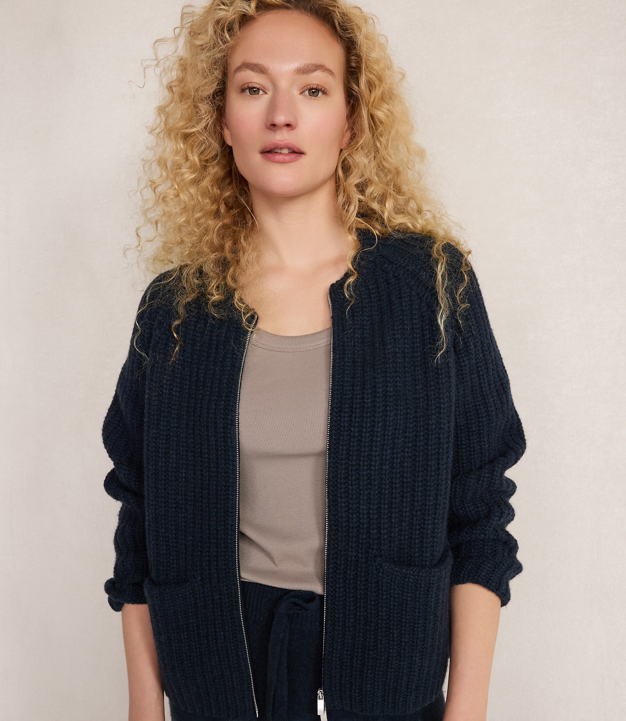 A woman with curly blonde hair wears a dark blue zippered cardigan over a taupe tank top, standing against a plain, light-colored background.