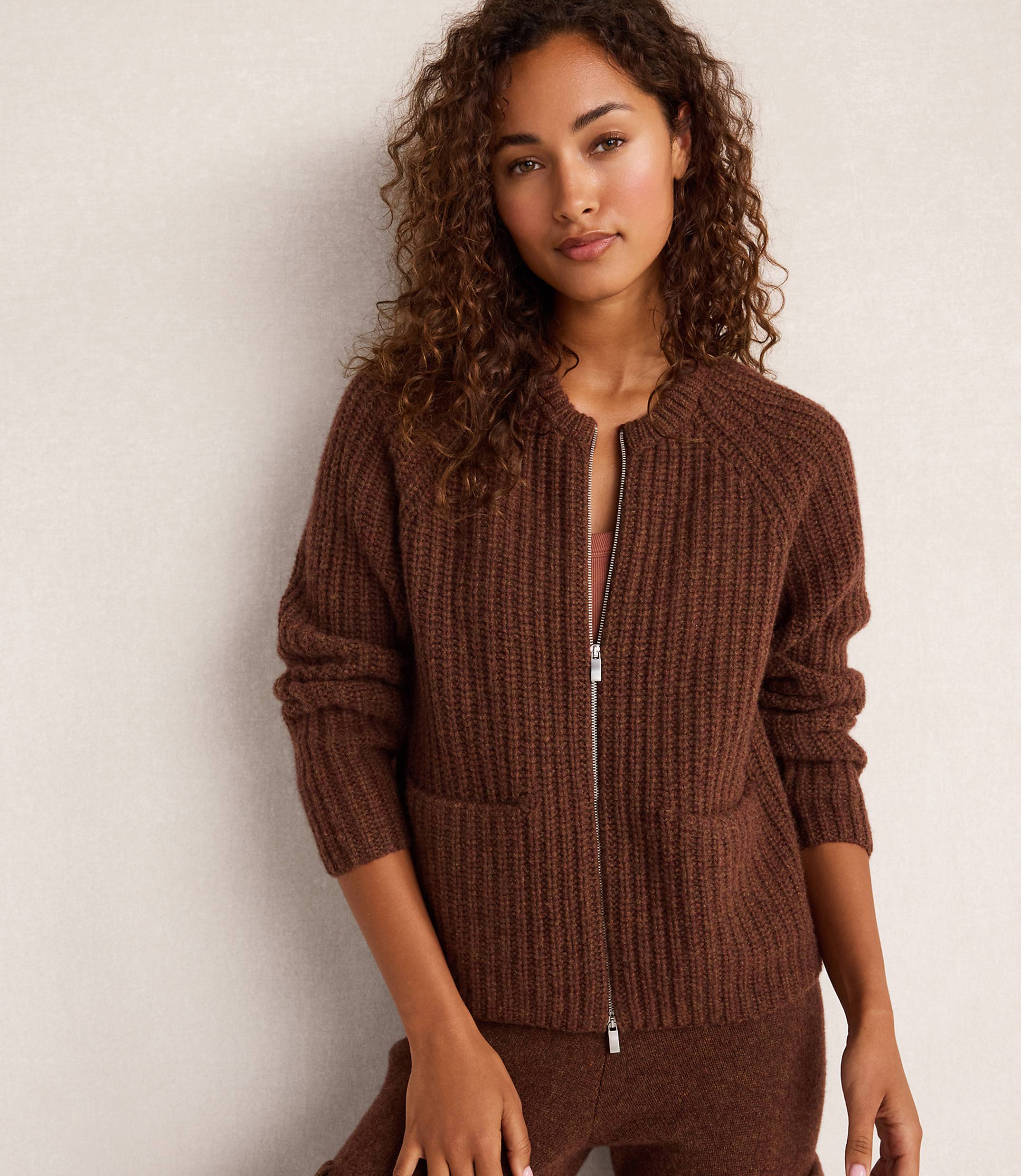 A woman with curly brown hair wears a brown ribbed zip-up cardigan with pockets, standing against a plain, light-colored wall.