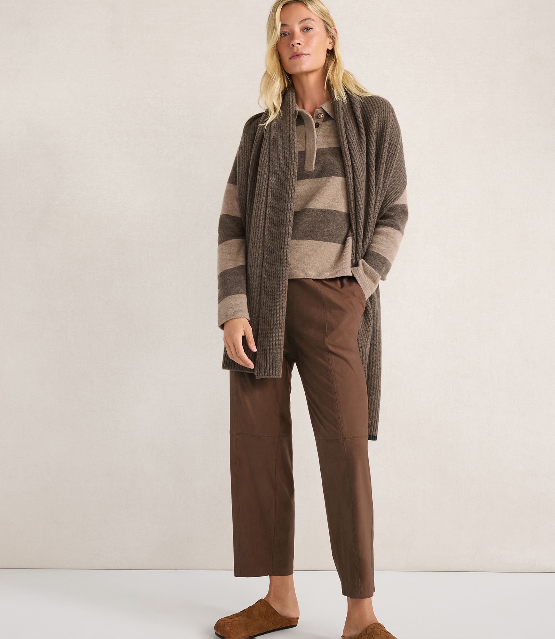 A woman in a brown striped sweater, brown pants, and a brown cardigan stands against a plain beige background.