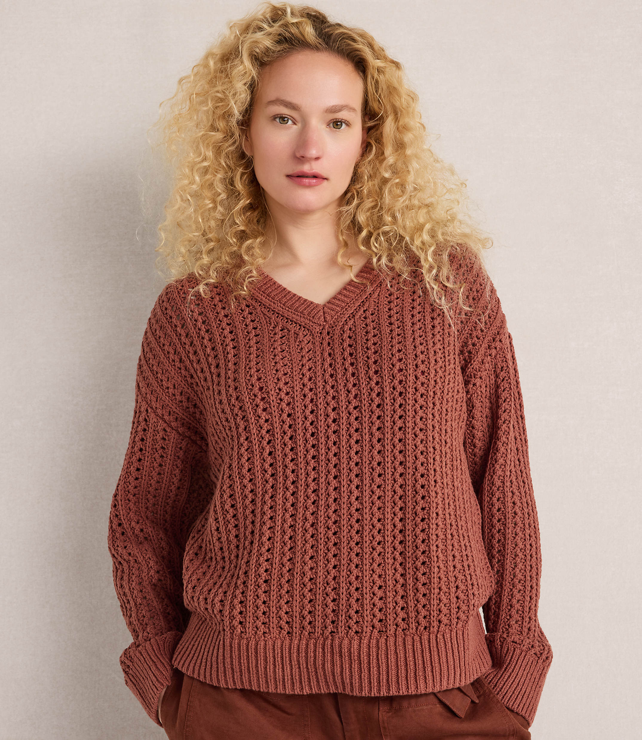 A woman with curly blonde hair wears a rust-colored v-neck sweater and brown pants against a plain, light-colored background.