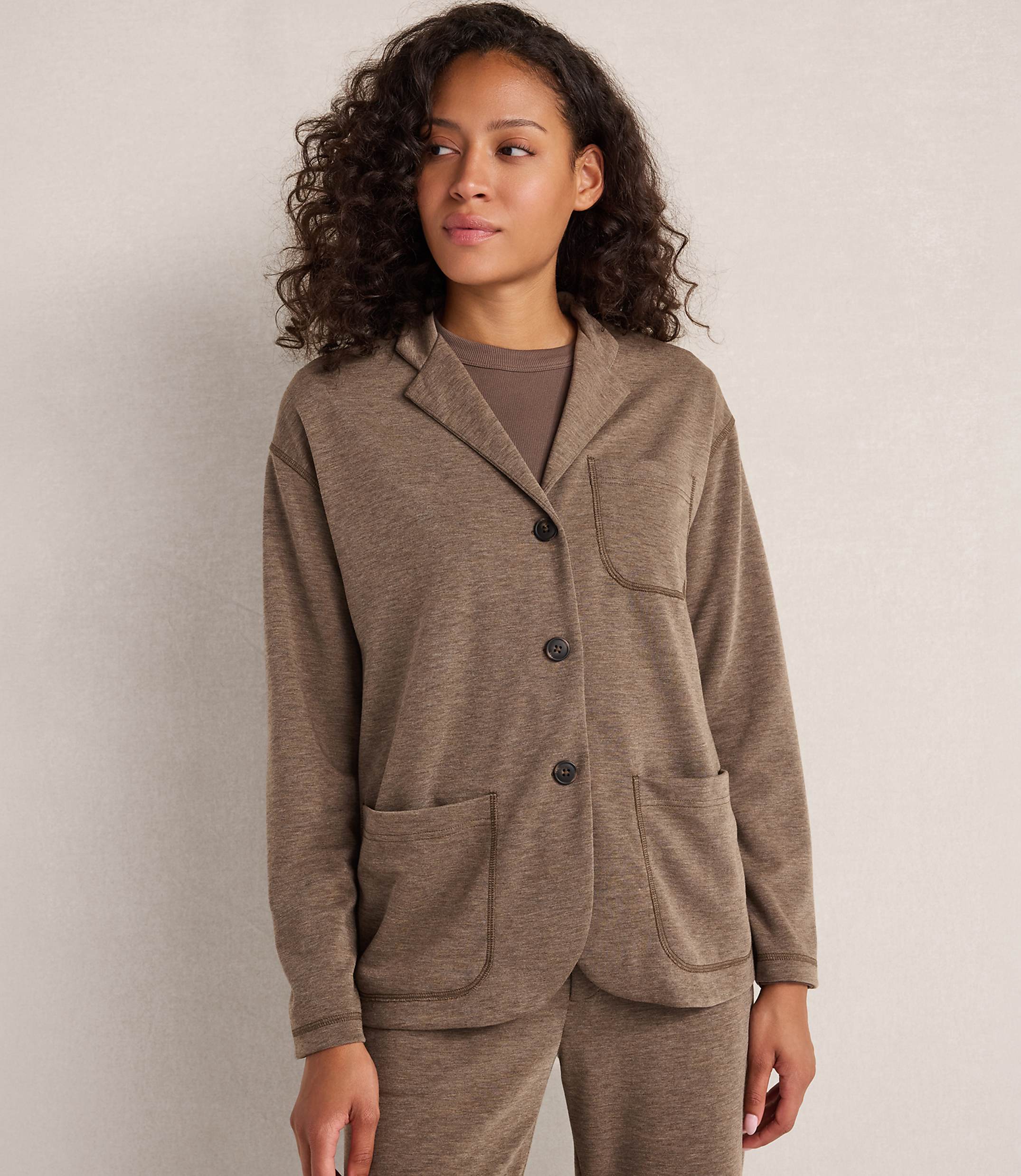 A woman with curly brown hair stands in the foreground wearing a brown blazer over a brown t-shirt and matching pants, against a plain, light-colored background.