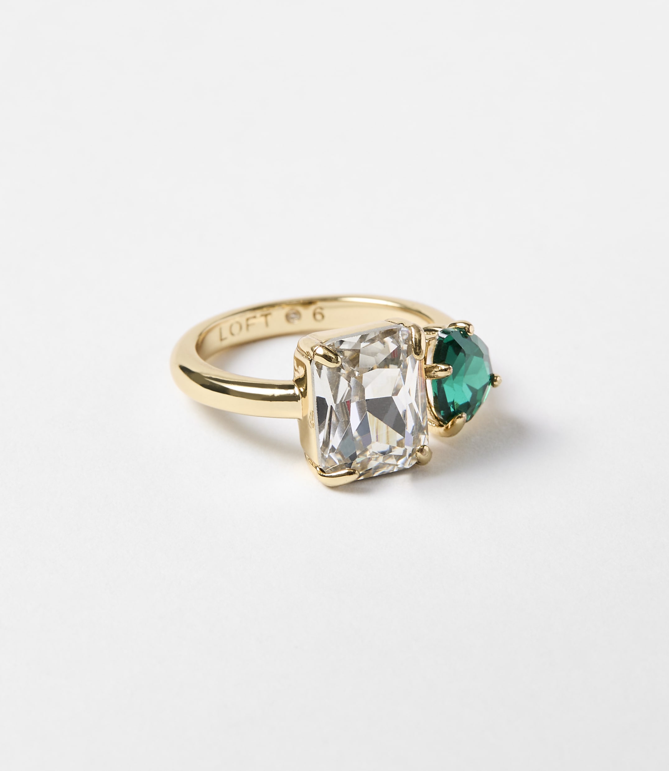 A gold ring with a rectangular clear stone and a heart-shaped green stone sits on a plain white background.