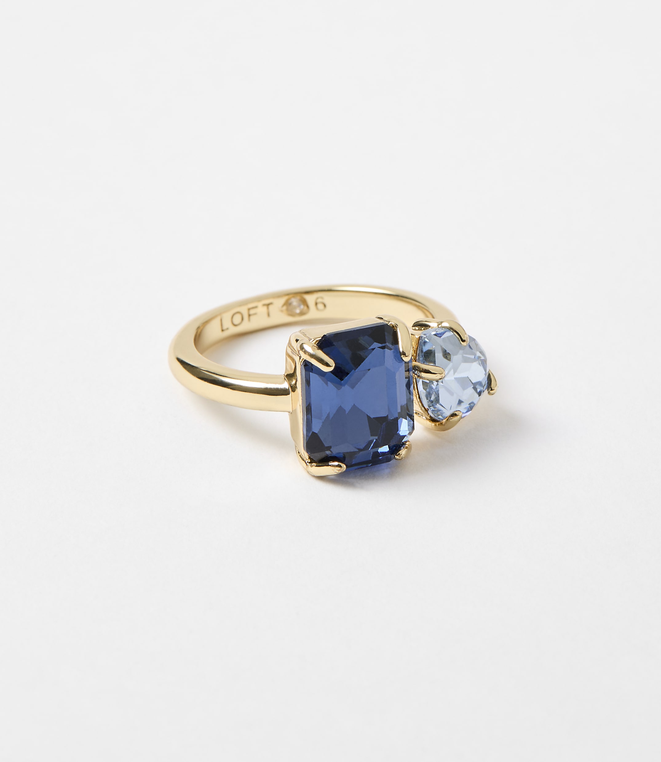 A gold ring with a large rectangular blue gemstone and a smaller heart-shaped light blue gemstone sits on a plain white background.
