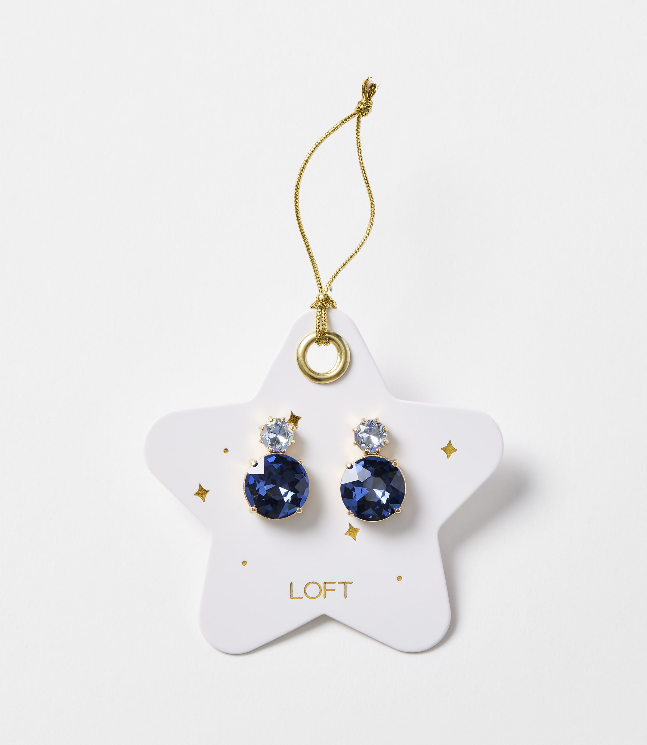 A pair of blue gem earrings are displayed on a white star-shaped card with gold accents and the "LOFT" logo, against a plain white background.