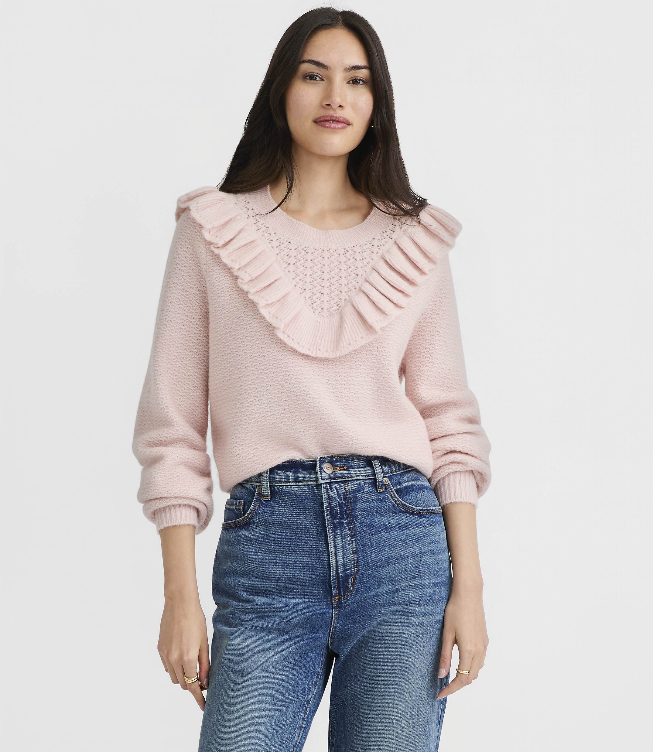A woman in a pink ruffled sweater and blue jeans stands against a plain white background.