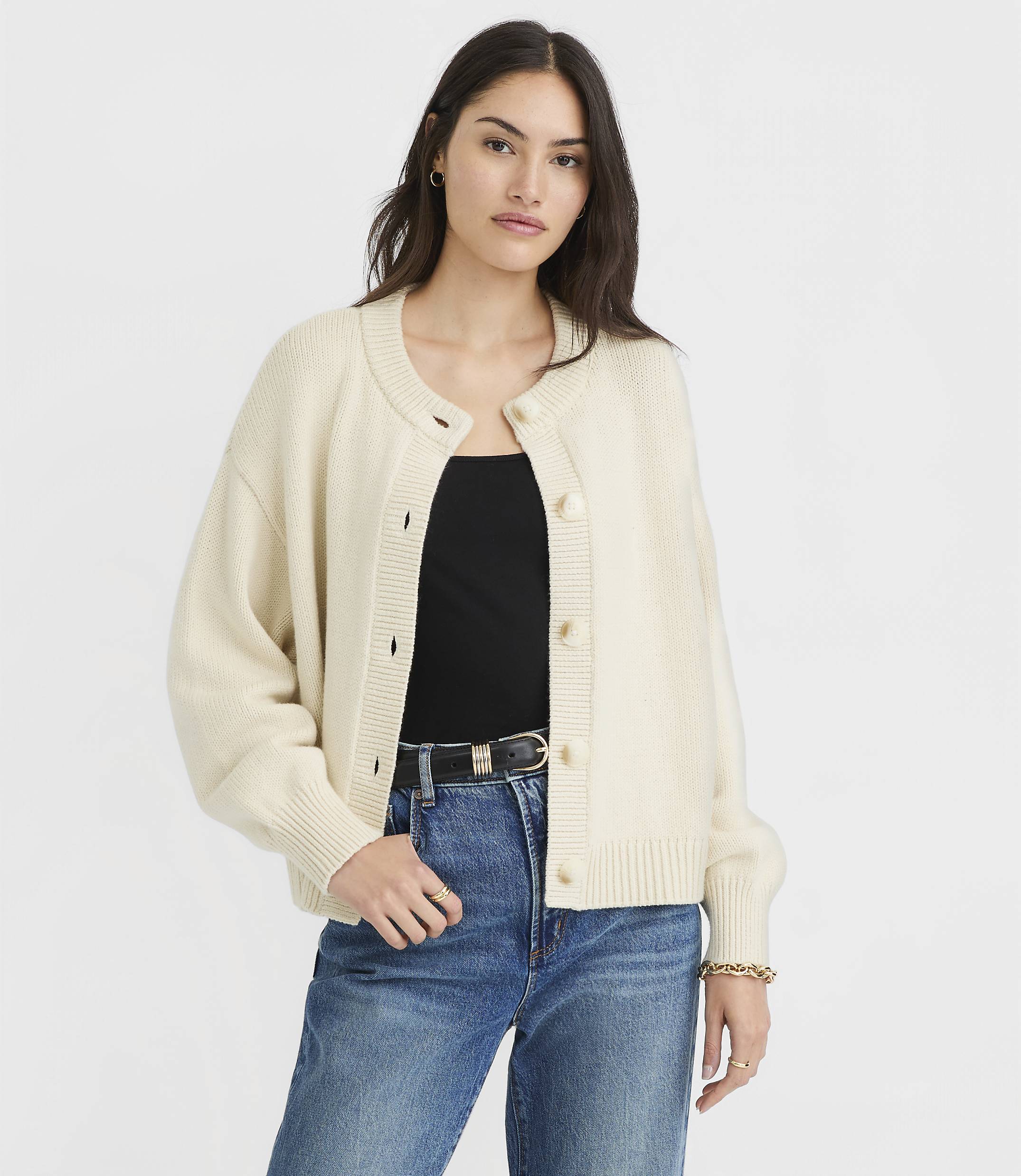 A woman in a cream cardigan and blue jeans stands against a plain white background.