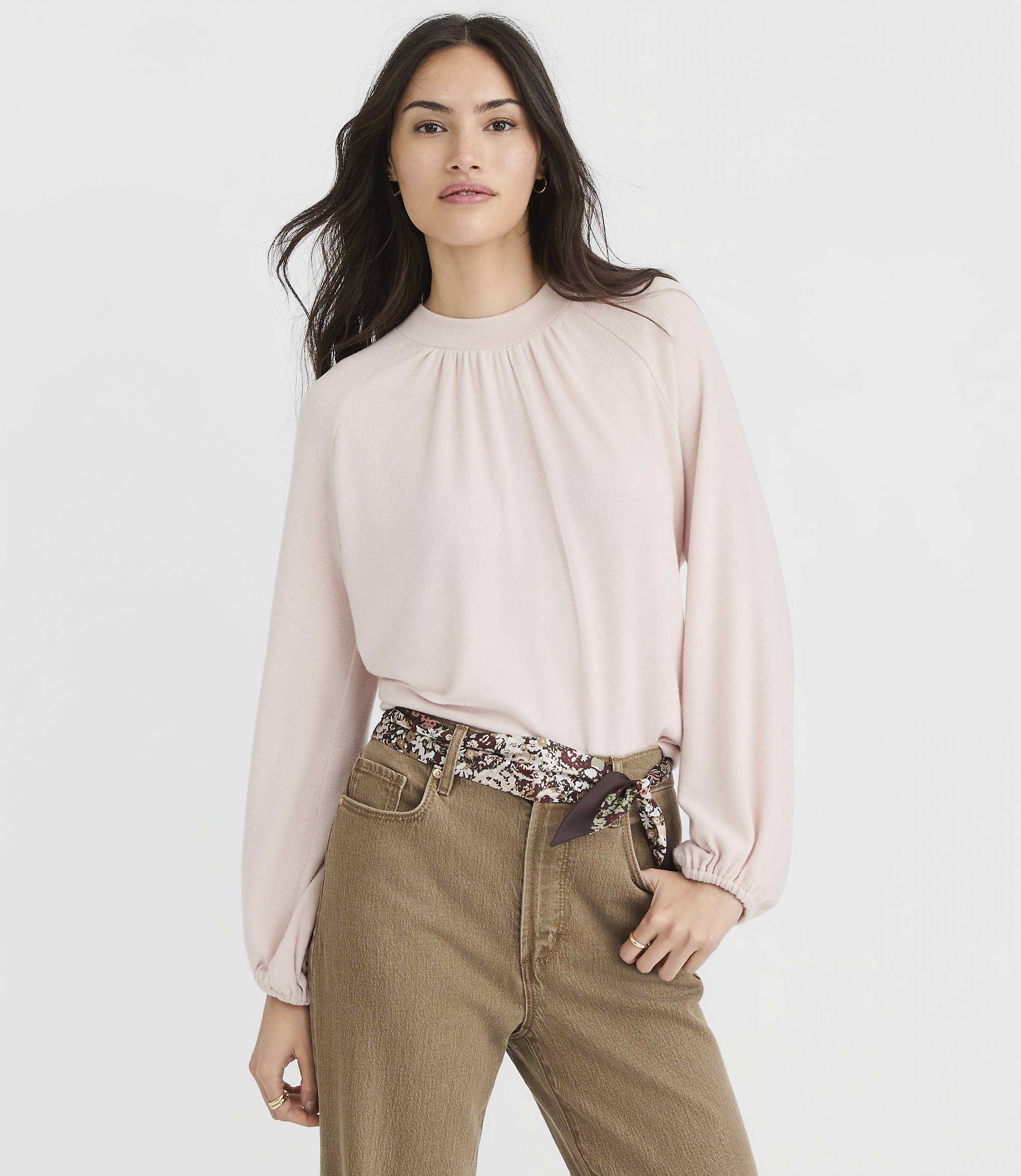 A woman in a pink blouse and brown pants stands against a plain gray background.