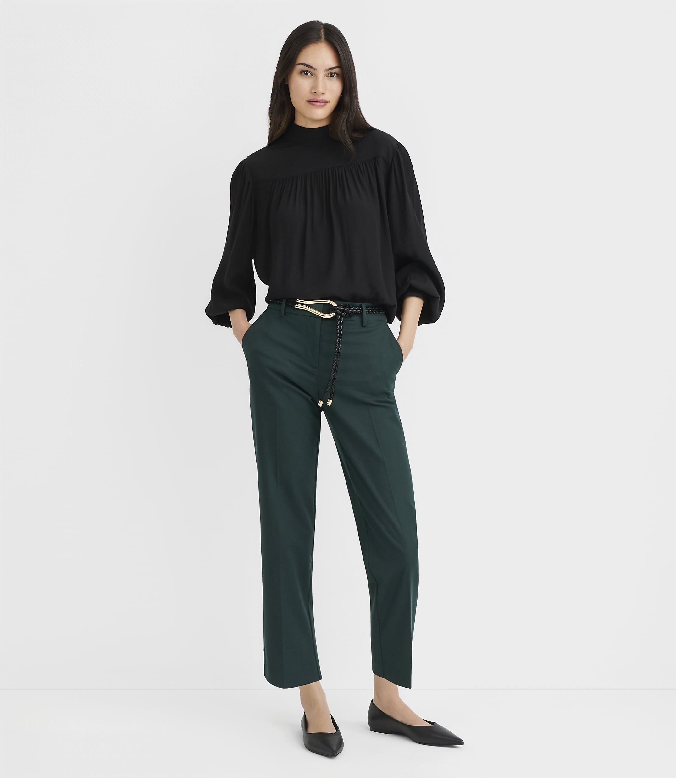 A woman in a black blouse and green pants stands against a plain white background.
