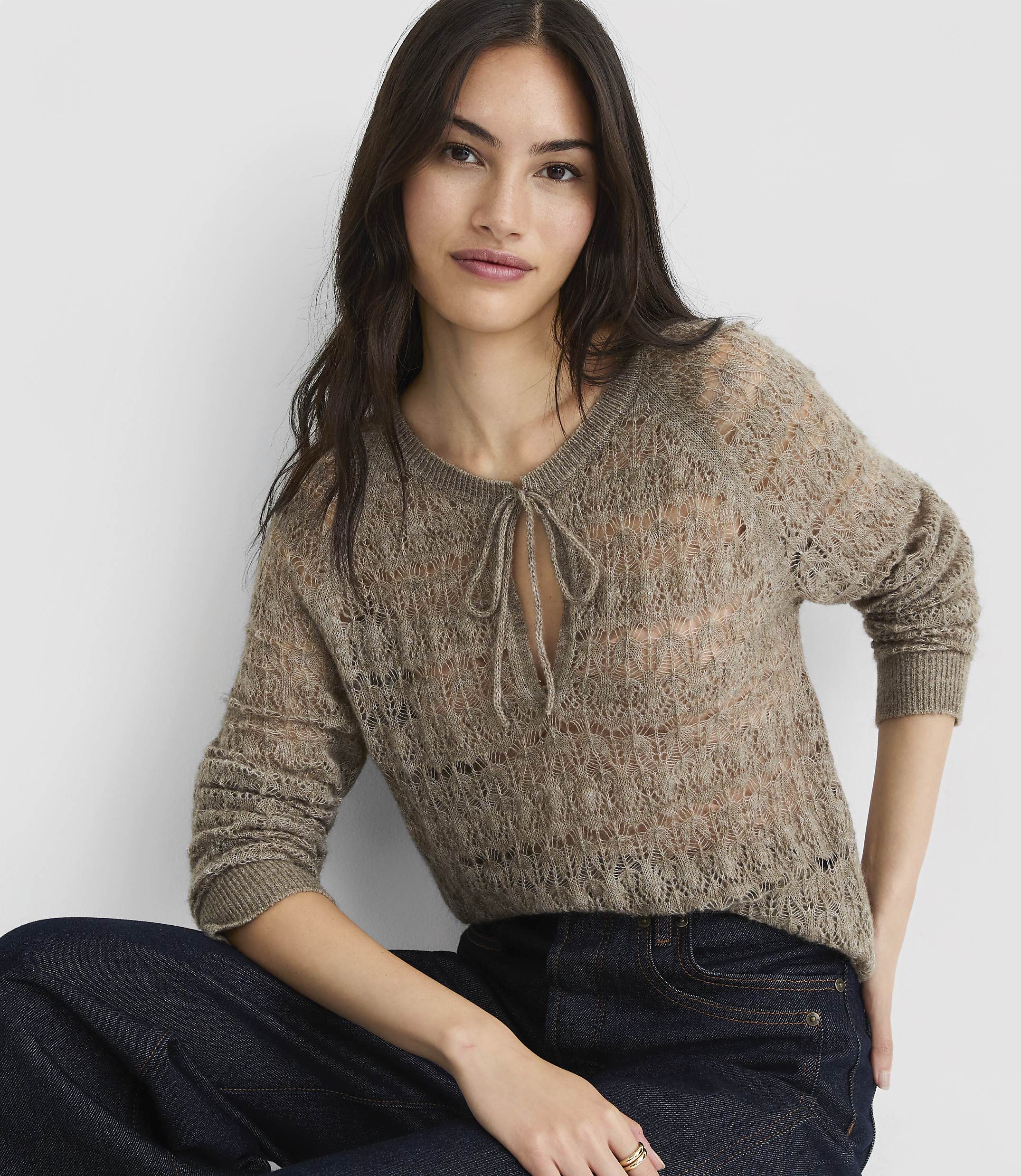 A woman in a sheer, taupe knit sweater with a tie-neck detail and dark wash jeans poses against a plain, light gray background.