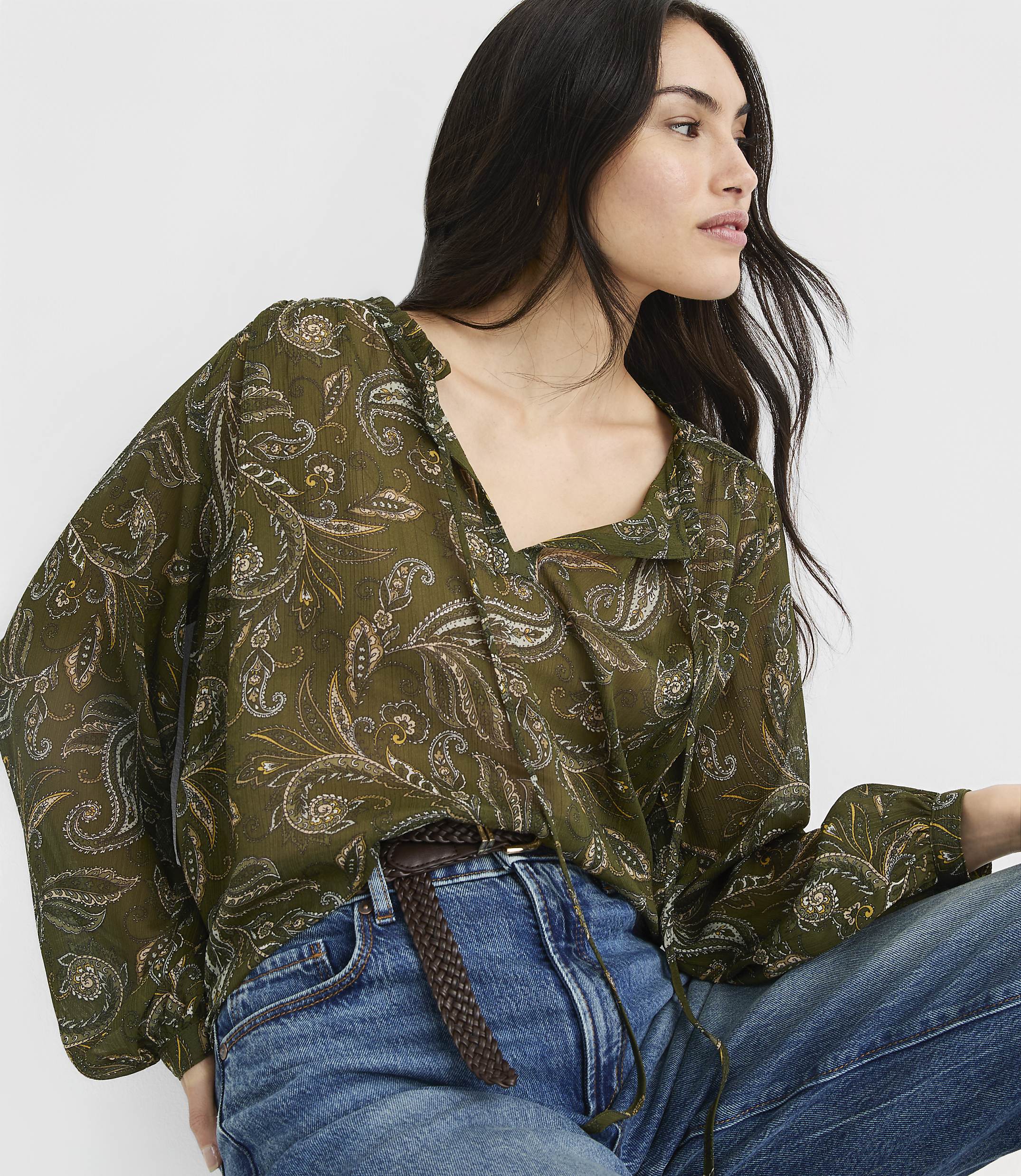 A woman in a flowing green paisley blouse and blue jeans is captured in a close-up shot against a plain white background.