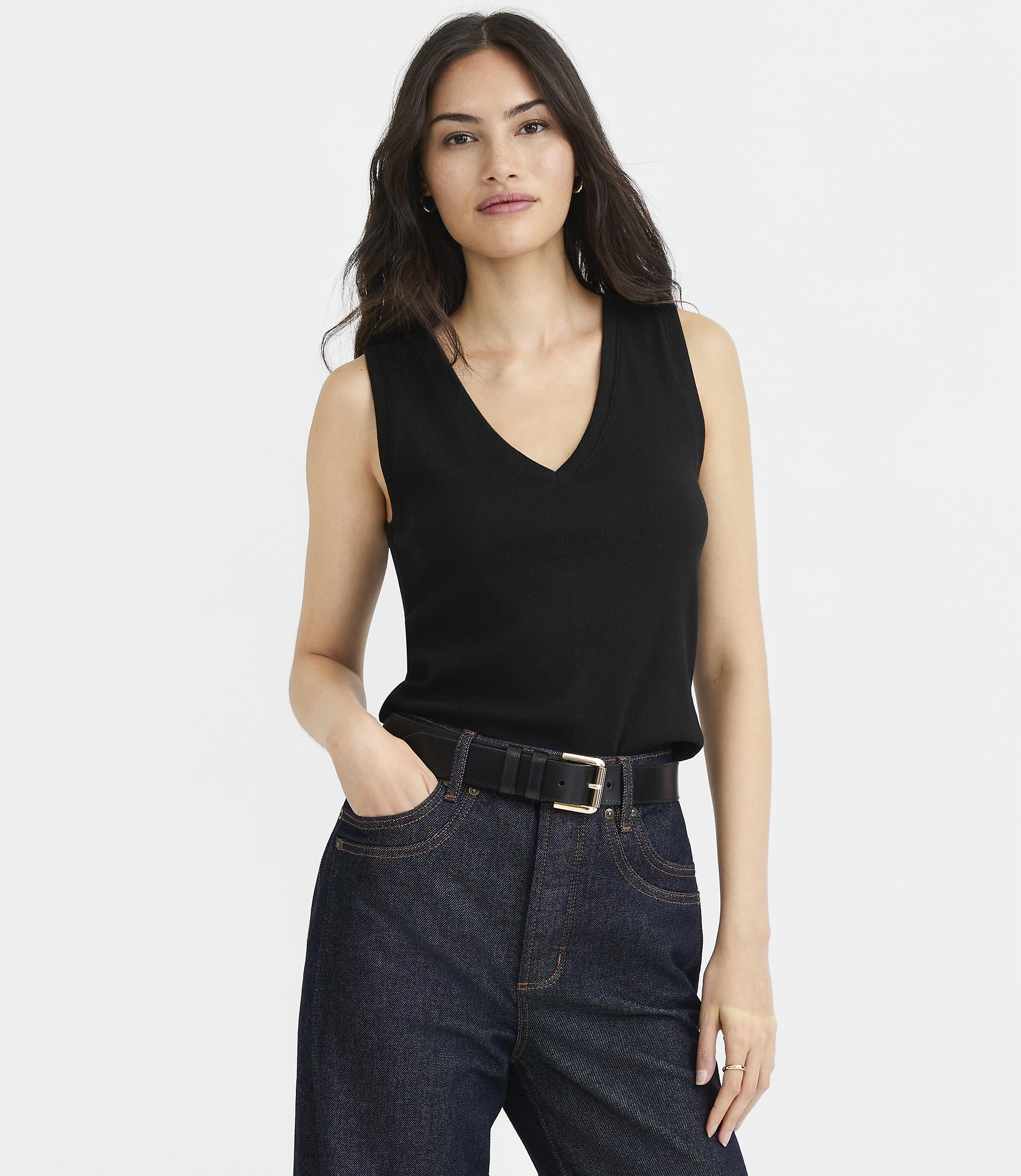 A woman in a black V-neck top and dark jeans stands against a plain white background.