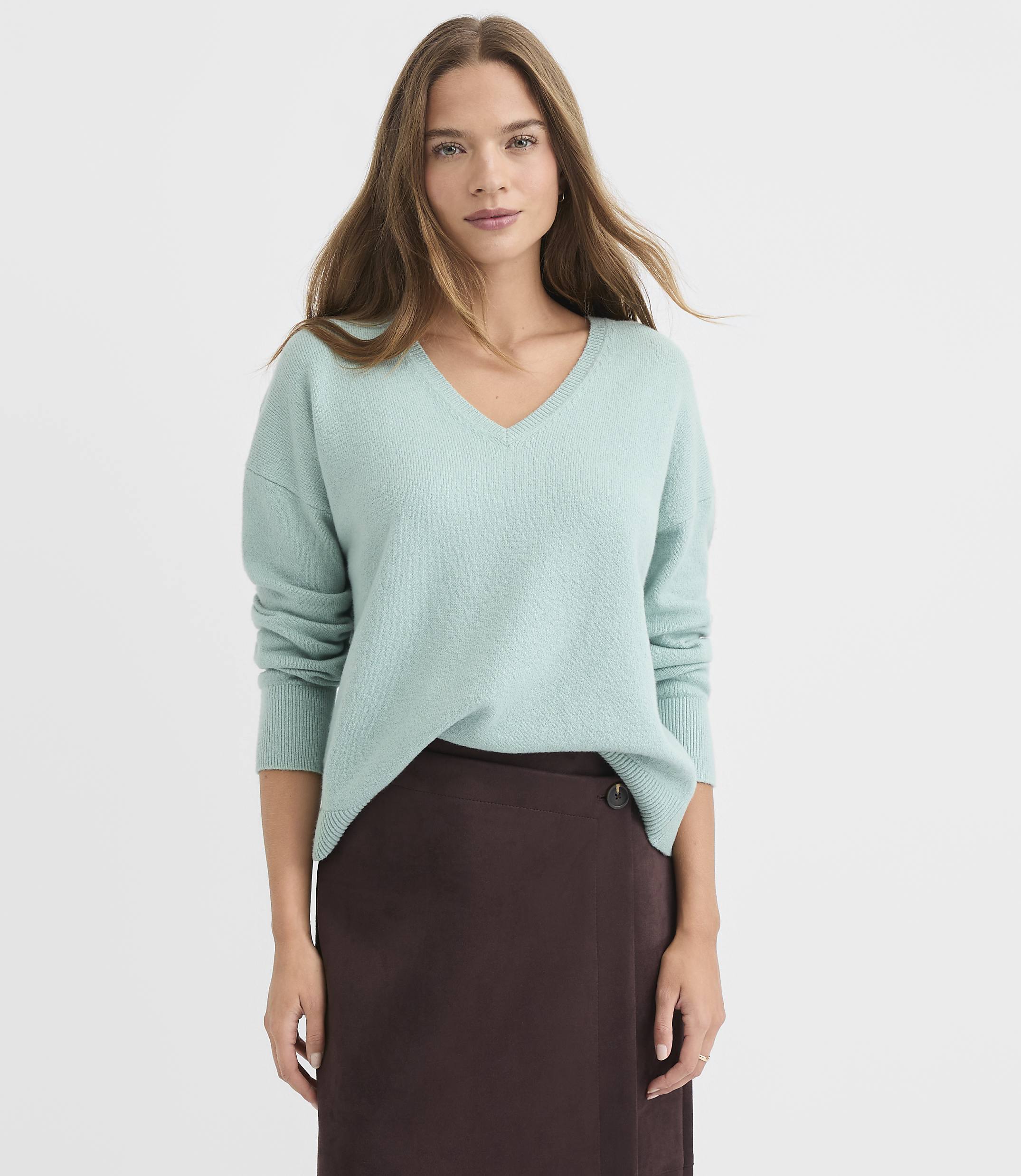 A woman in a light blue v-neck sweater and a brown skirt stands against a plain white background.