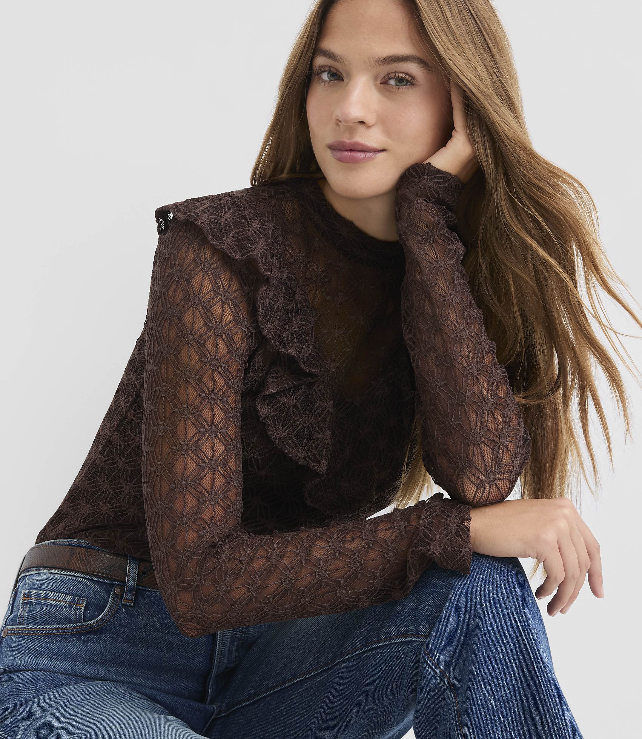 A woman in a brown lace top and blue jeans poses against a plain white background.