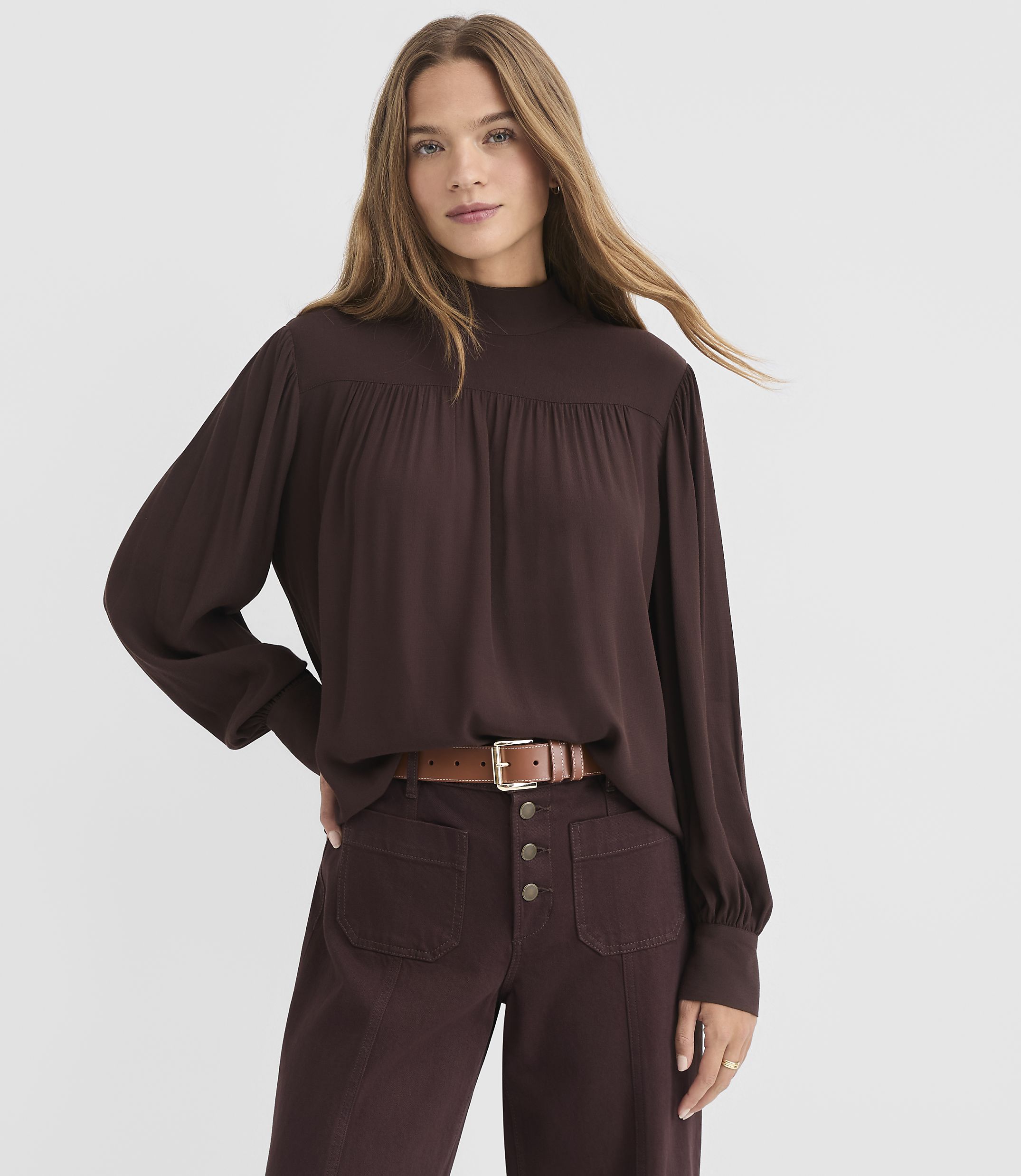 A woman in a brown mock-neck blouse and maroon pants stands against a plain white background.