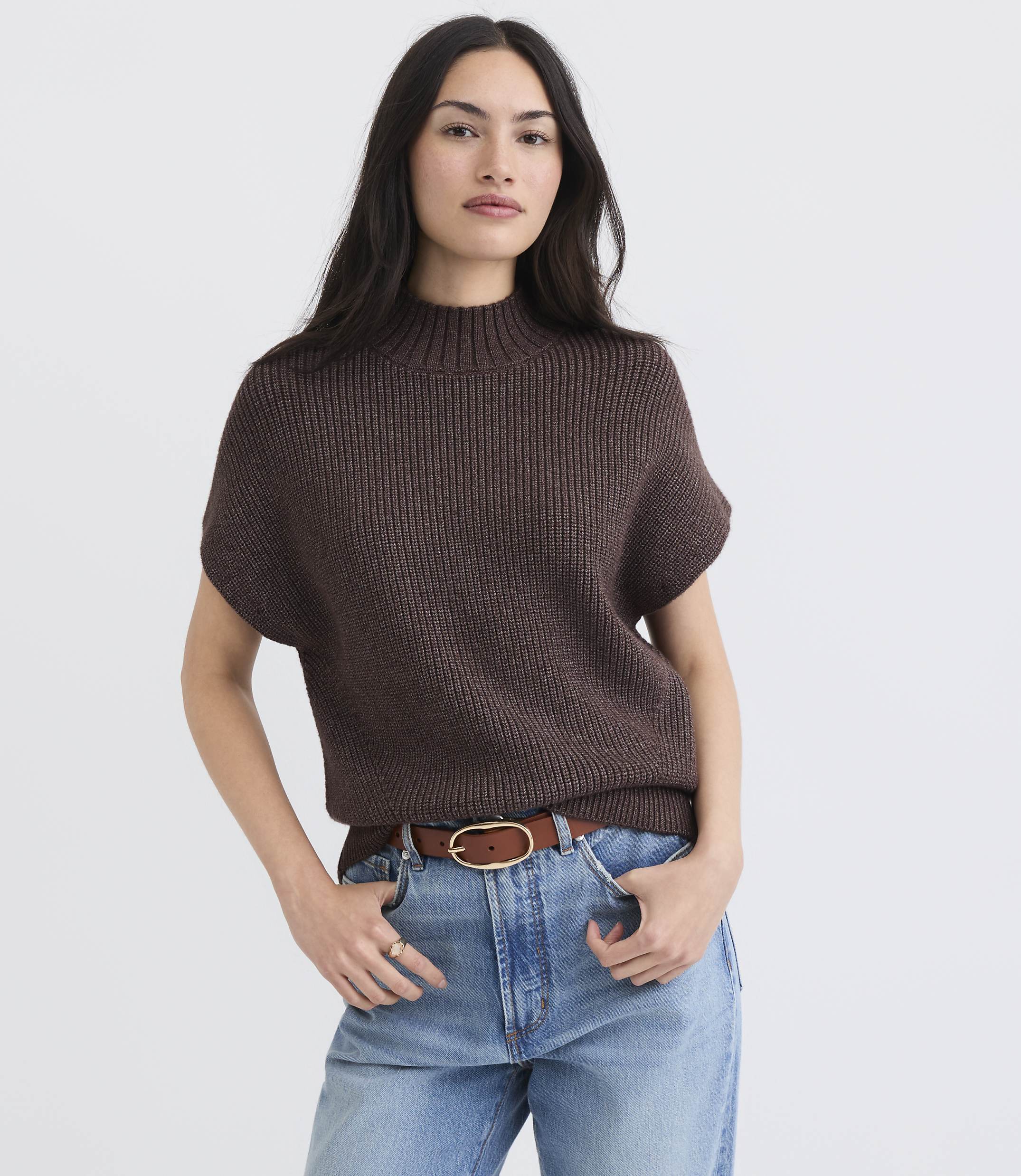 A woman in a brown mock-neck sweater and blue jeans stands against a plain white background.