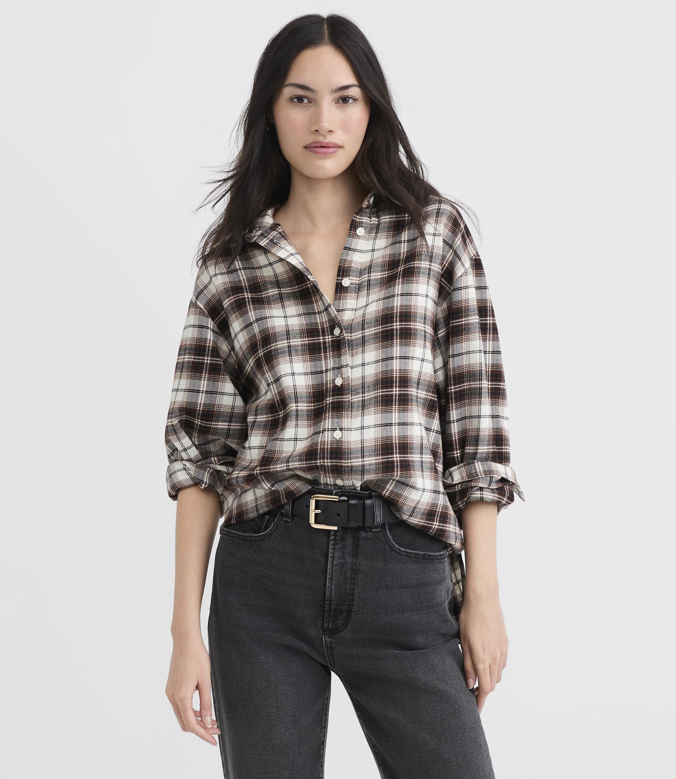 A woman in a brown plaid shirt and black jeans stands against a plain white background.