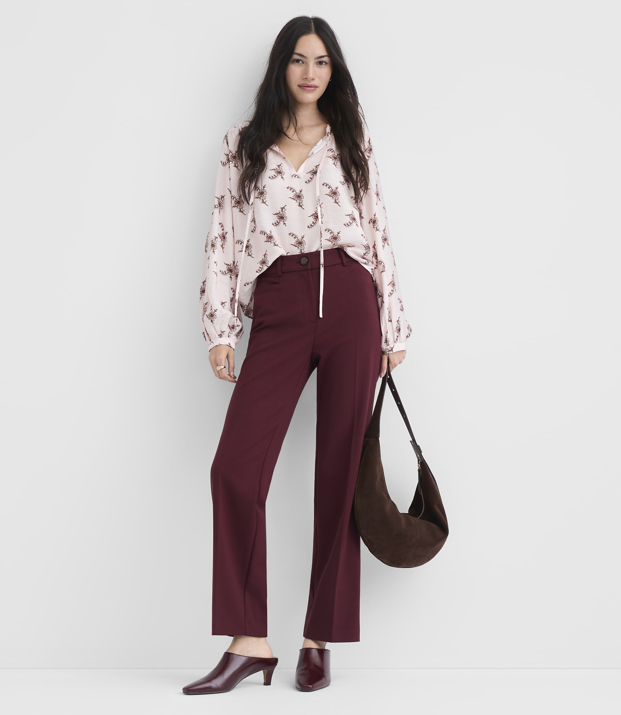 A woman in a floral blouse and maroon pants stands against a plain white background, holding a brown suede purse.