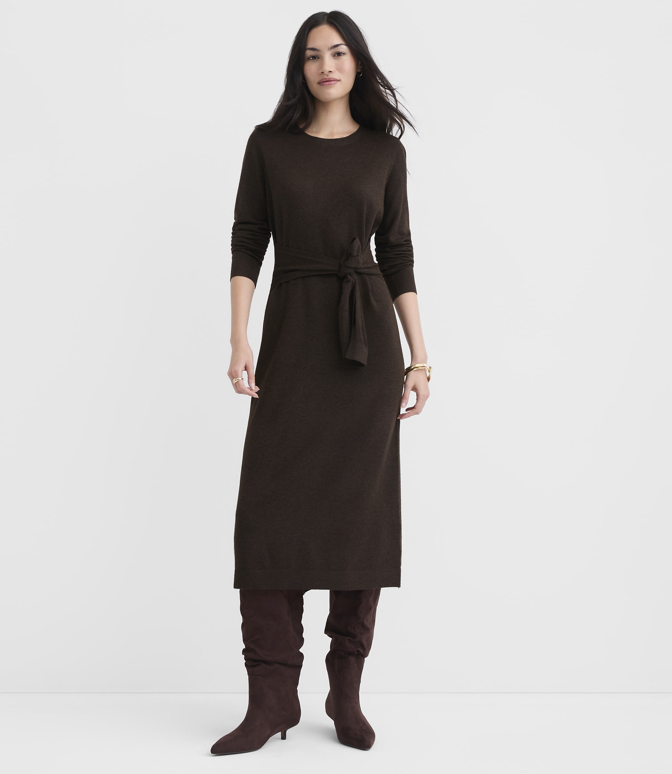 A woman in a brown knit dress and boots stands against a plain white background.
