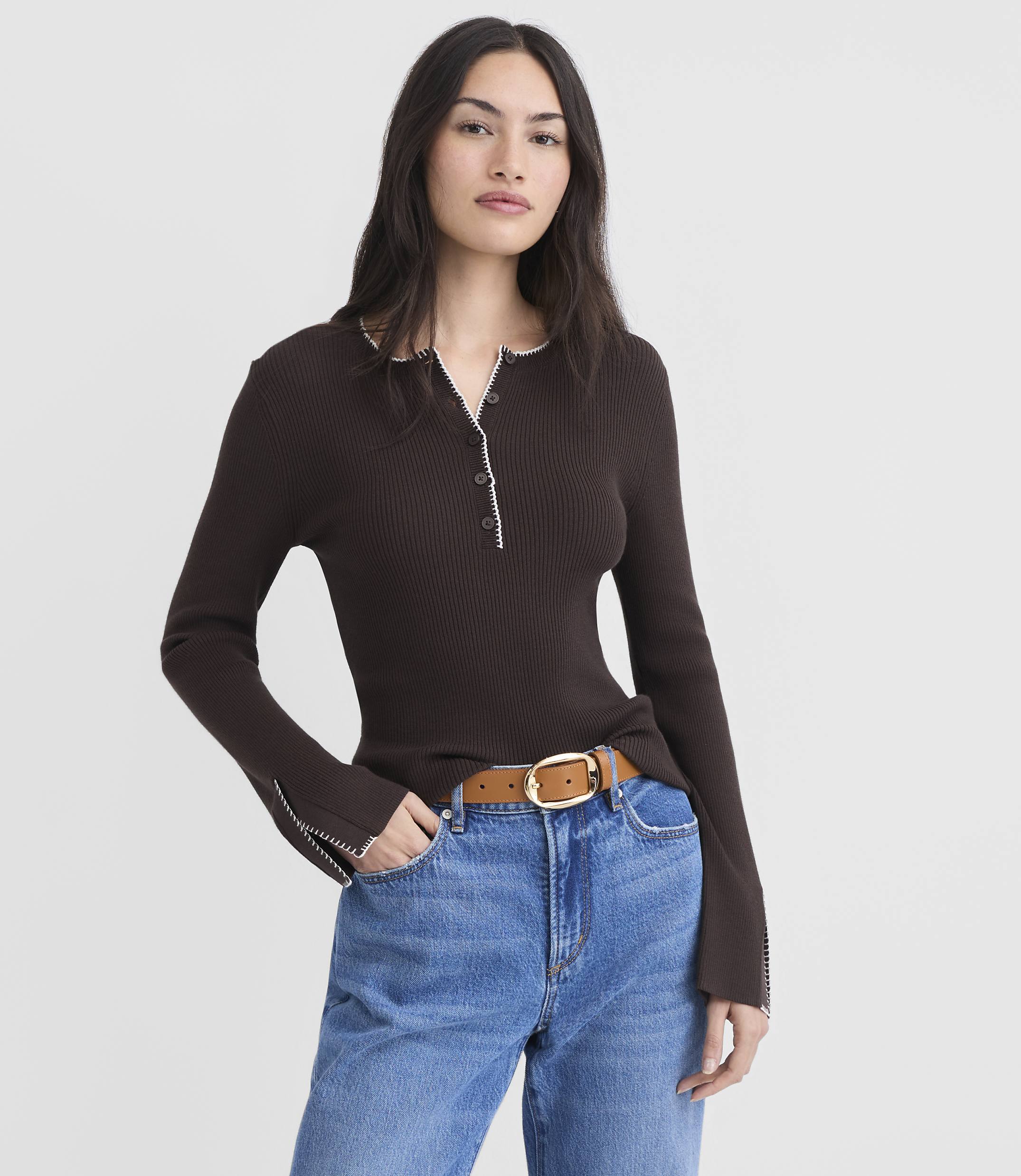 A woman in a brown ribbed shirt and blue jeans stands against a plain white background.