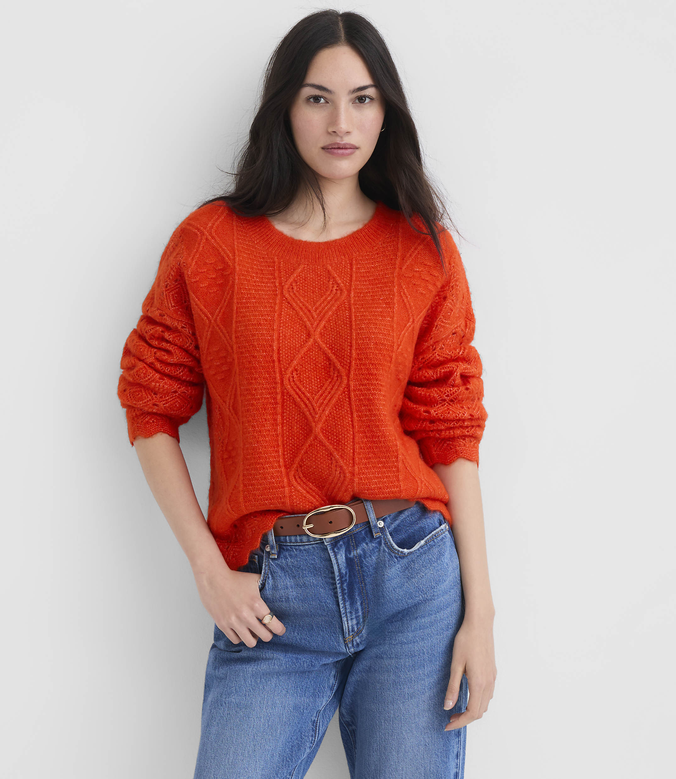 A woman in a bright orange cable-knit sweater and blue jeans stands against a plain white background.