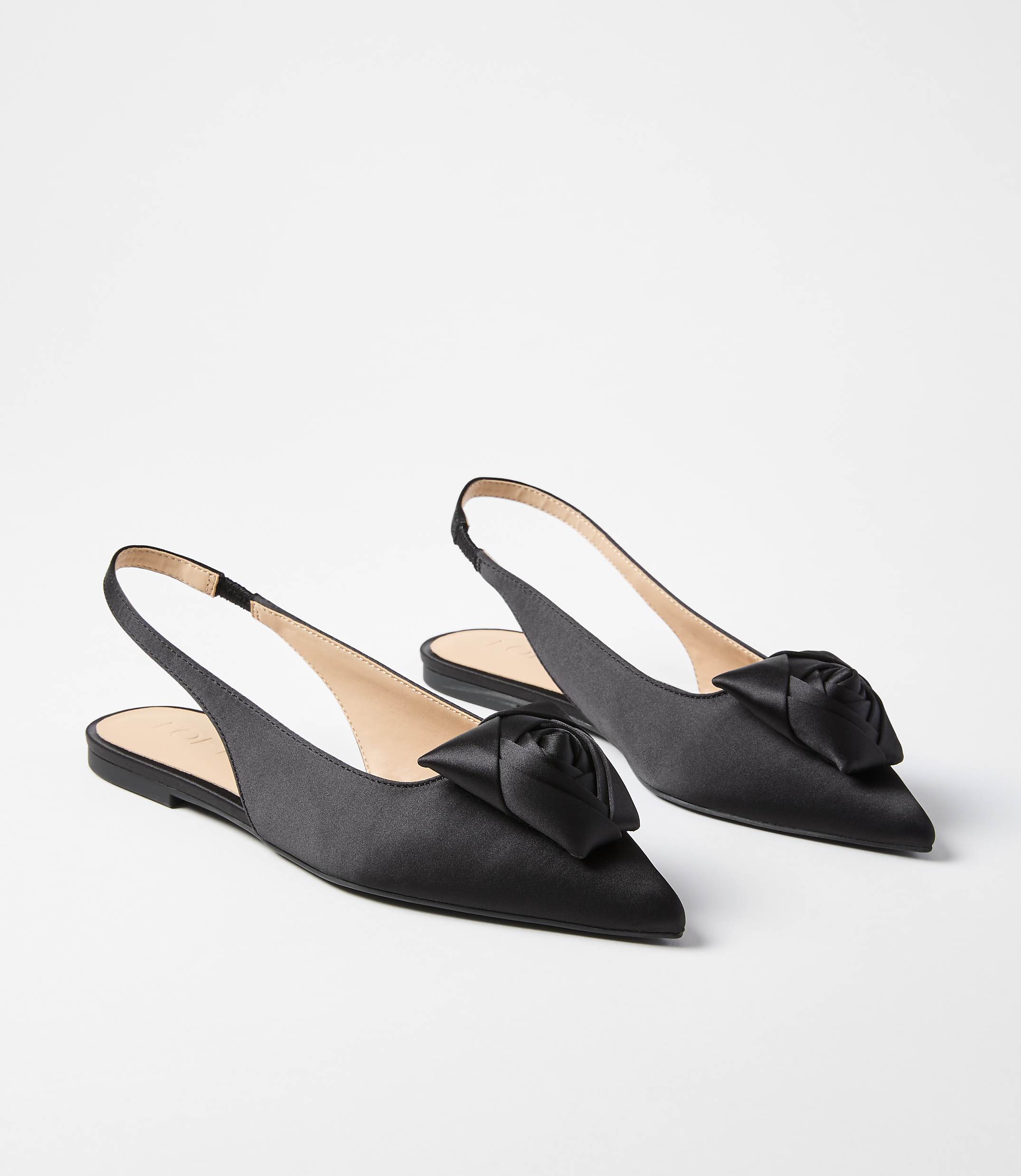 A pair of black satin slingback flats with a rose embellishment on the toe rest on a plain white background.
