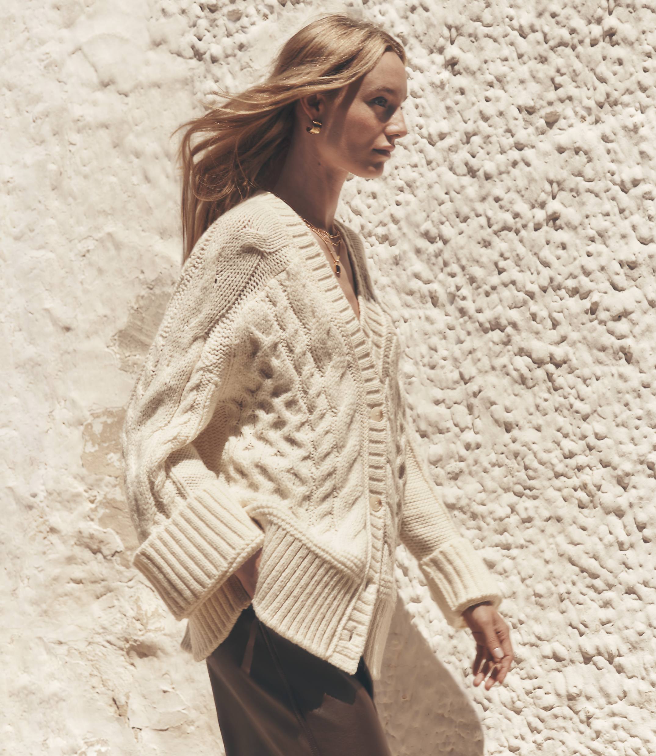A blonde woman in a cream cable-knit cardigan walks against a textured, light-colored wall.