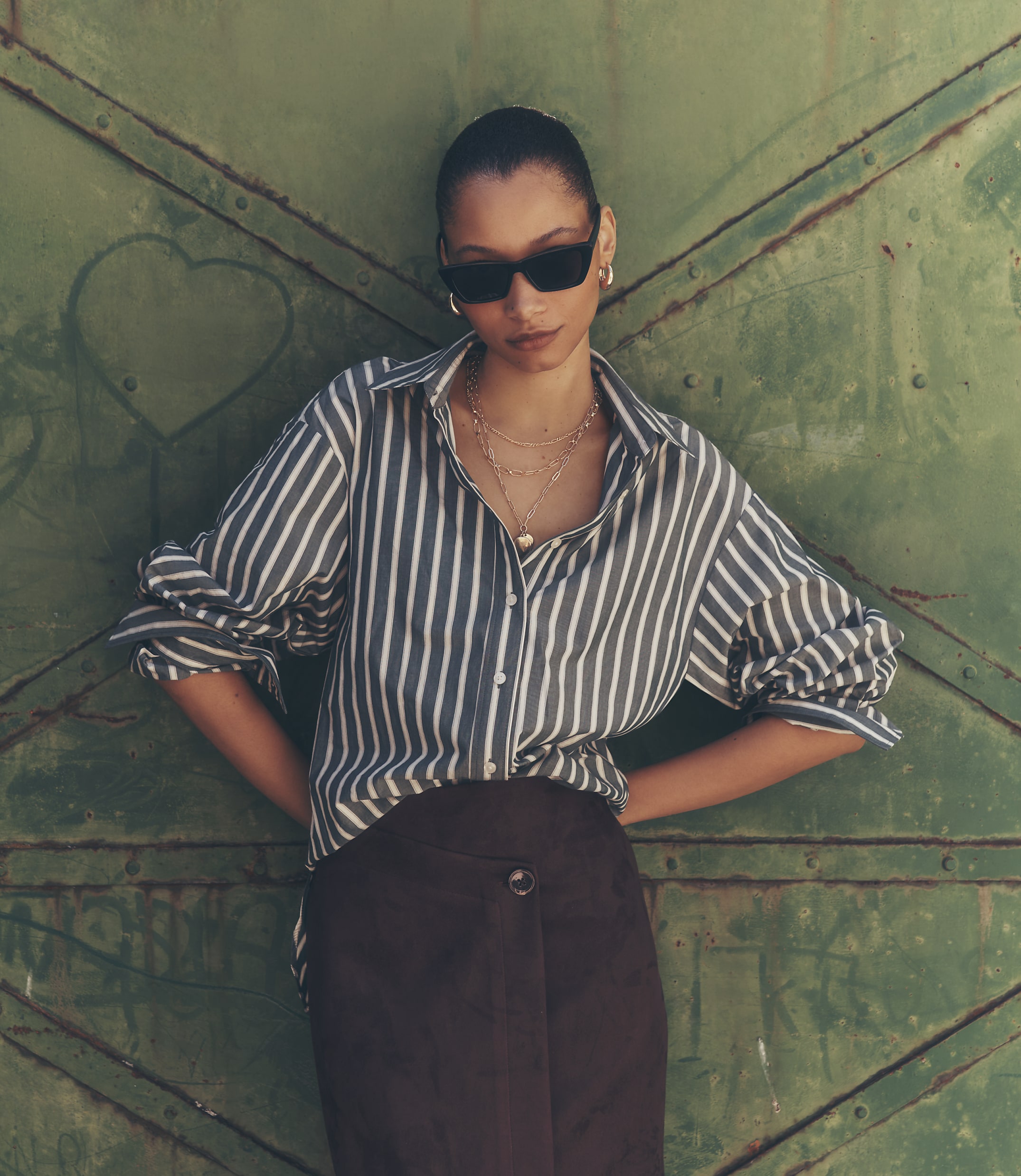 A woman in a striped shirt and dark skirt stands against a green, textured metal door.