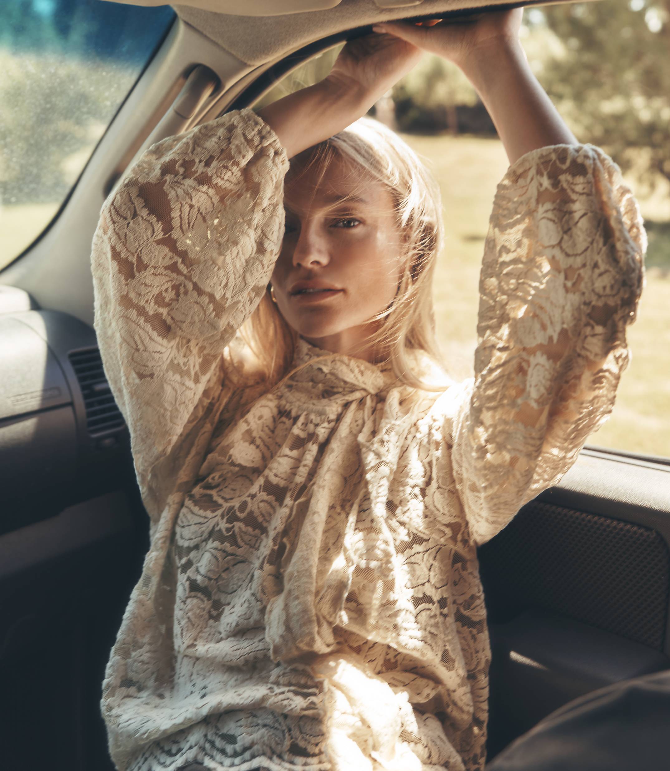 A woman in a lace top leans back in a car, her arms raised to the roof, with a sunlit, grassy landscape visible through the window.