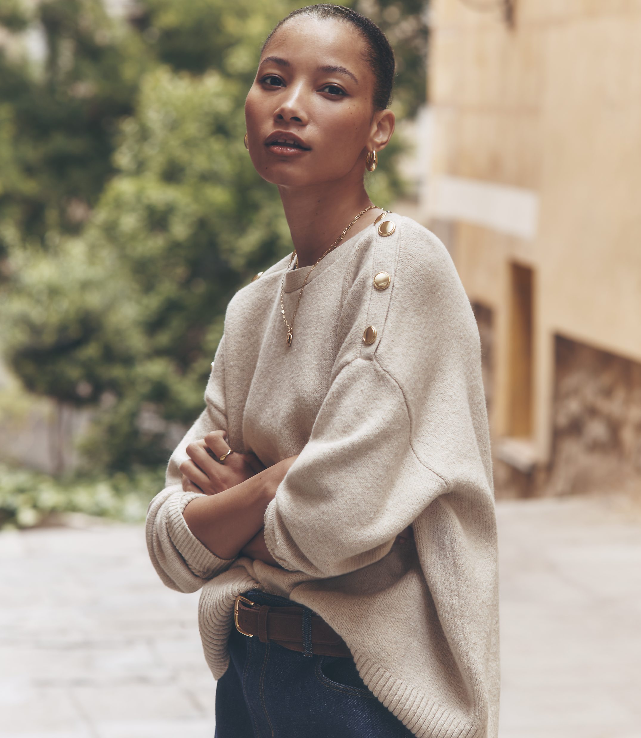 A woman in a cream-colored sweater with gold buttons stands with her arms crossed in front of a blurred background of a European street.