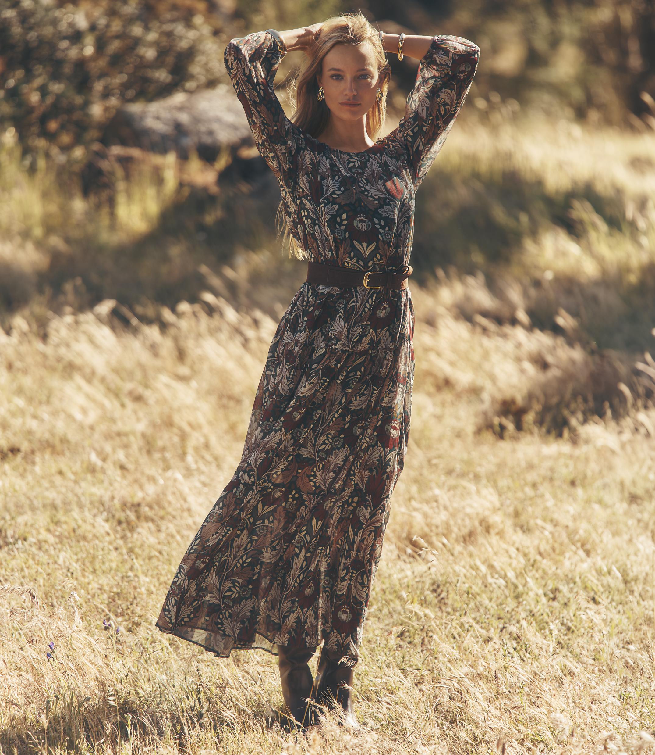 A woman in a floral dress stands in a dry, grassy field with her hands behind her head, while a blurred, sunlit landscape stretches out behind her.